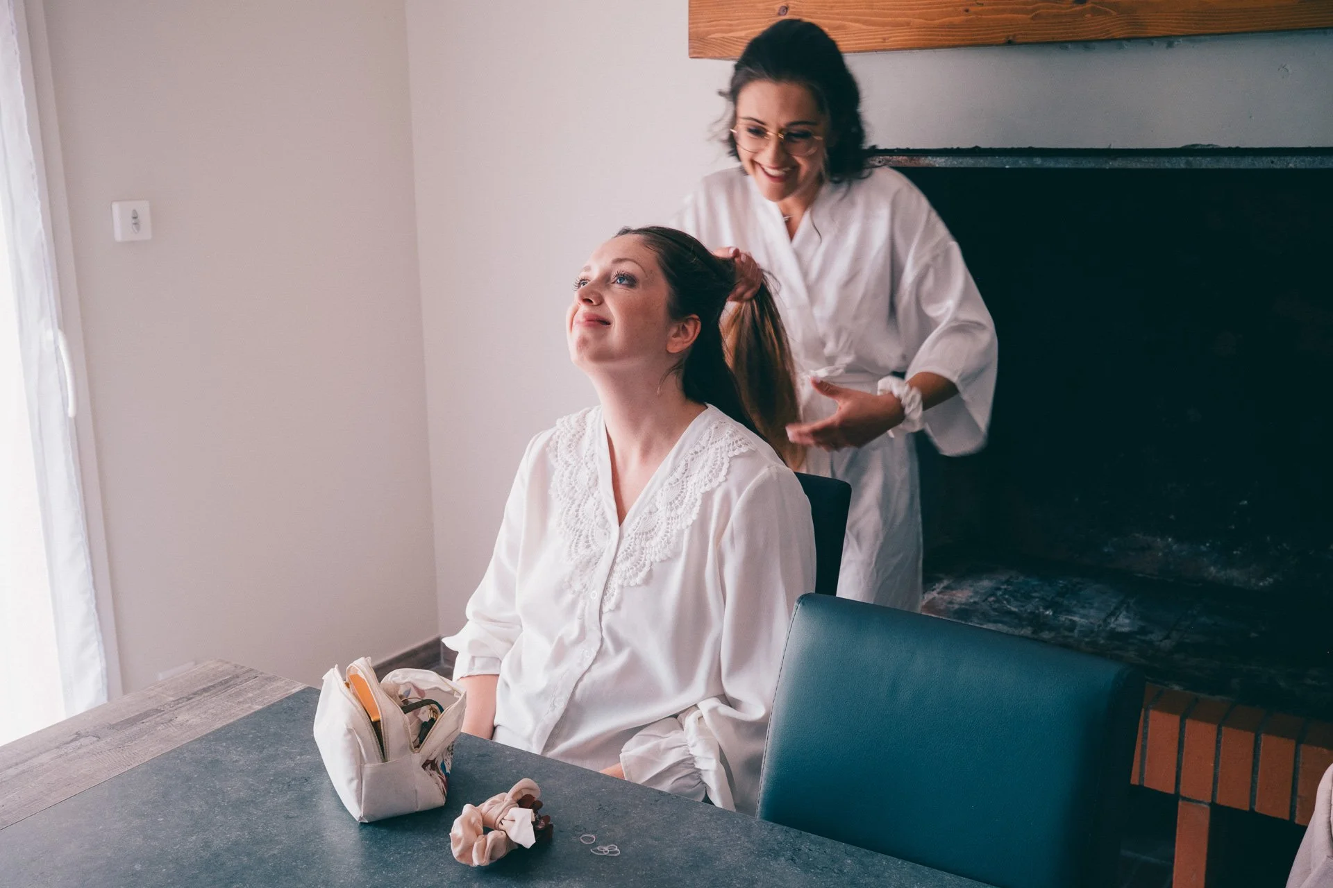 Une femme est assise à une table, une autre femme lui fait une coiffure. La femme assise porte une blouse blanche, un sac à main coloré est sur la table.