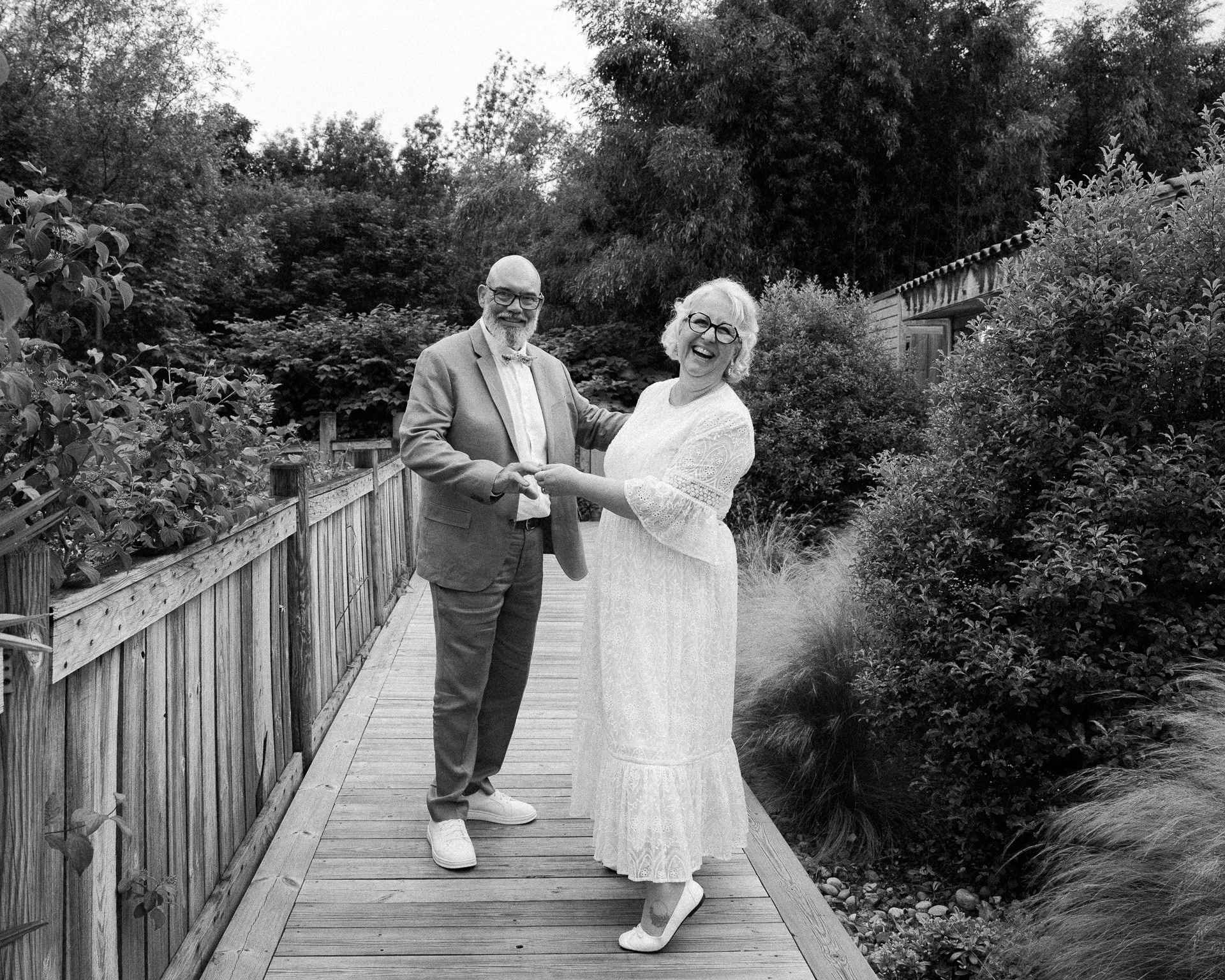 Un homme et une femme âgés dans un jardin, se tenant par la main et riant, vêtus de tenues élégantes, en train de partager un moment de bonheur