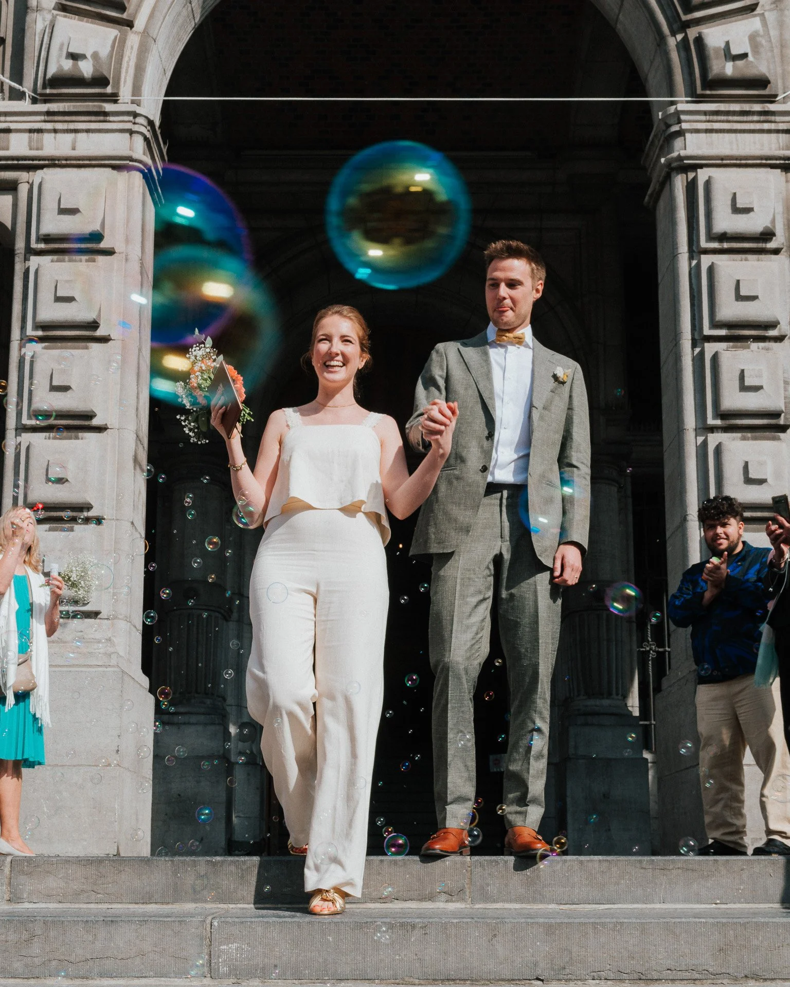 Un couple de mariés heureux marche main dans la main devant un bâtiment historique, entouré de proches, avec des bulles de savon flottant autour.