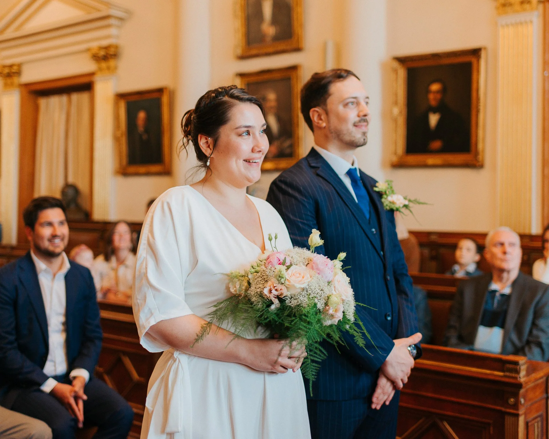 Un couple de mariés à une cérémonie religieuse, la femme tient un bouquet de fleurs et porte une robe blanche, l'homme porte un costume sombre avec une boutonnière, en arrière-plan des invités assis, dans une salle richement décorée avec des portrait