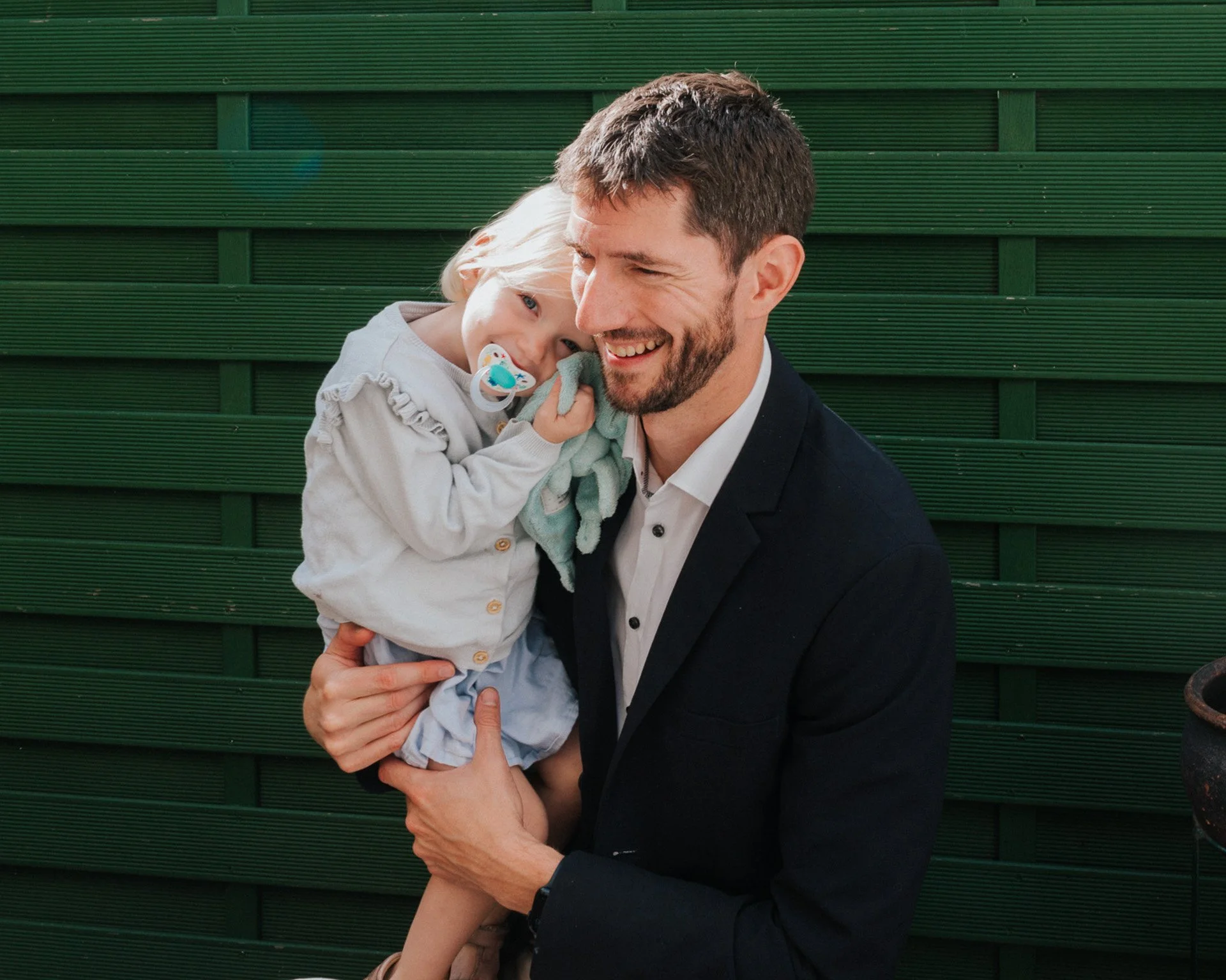 Un homme souriant tient une petite fille dans ses bras devant un mur vert. La petite fille a un tétine et porte une chemise beige avec des détails en volants.