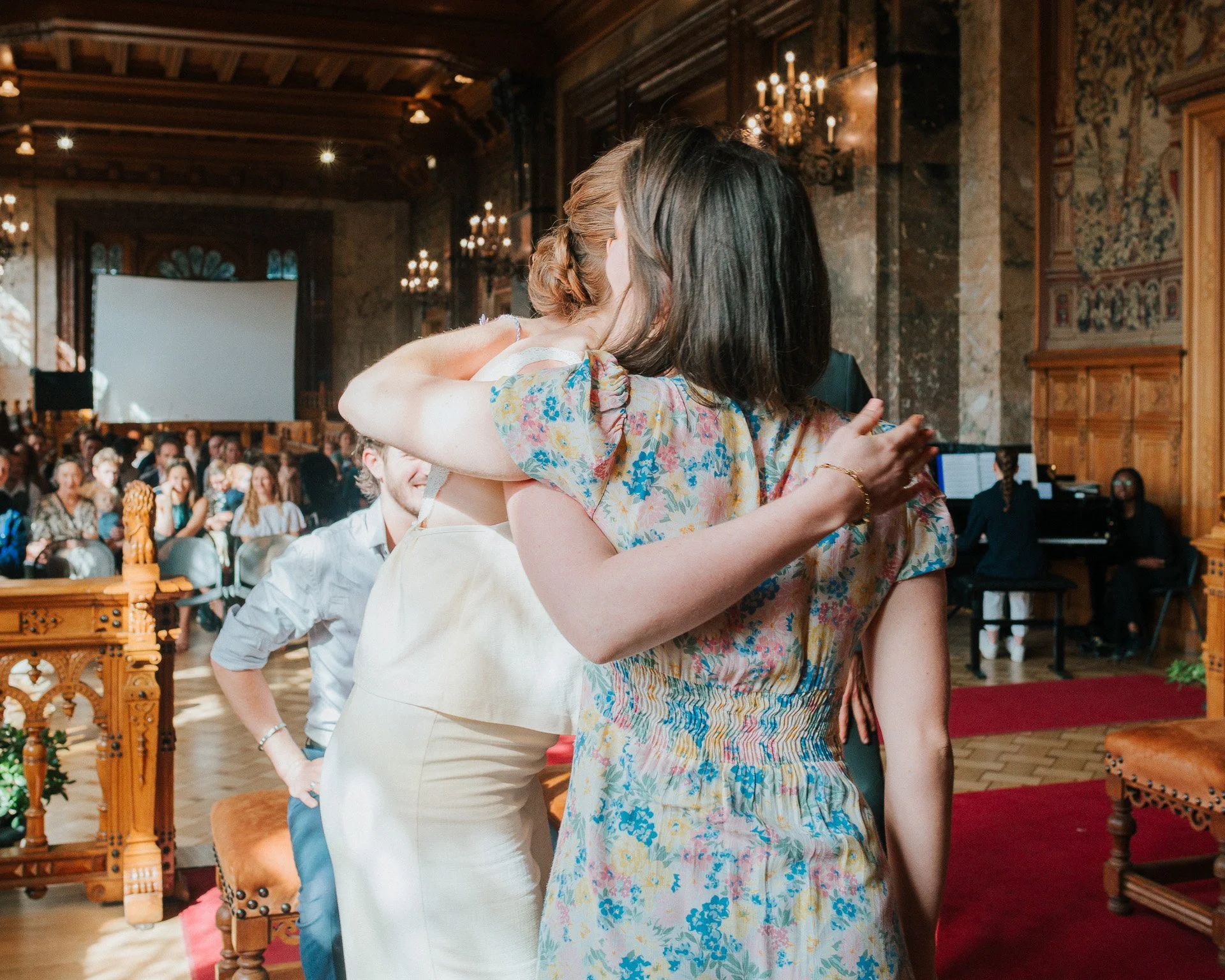 Deux femmes s'embrassent en plein air dans une salle de réception élégante avec des chandeliers et une foule assise en arrière-plan.