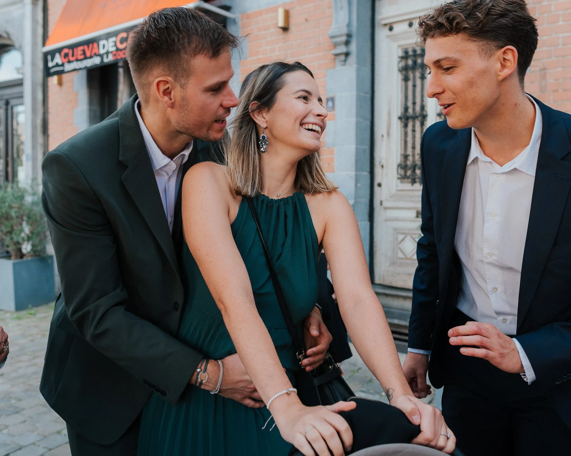 Trois jeunes adultes, deux hommes et une femme, souriants et en costume, discutent et rient à l'extérieur devant un bâtiment en briques, dans un cadre urbain. La femme porte une robe verte et des boucles d'oreilles, le groupe semble convivial et déte