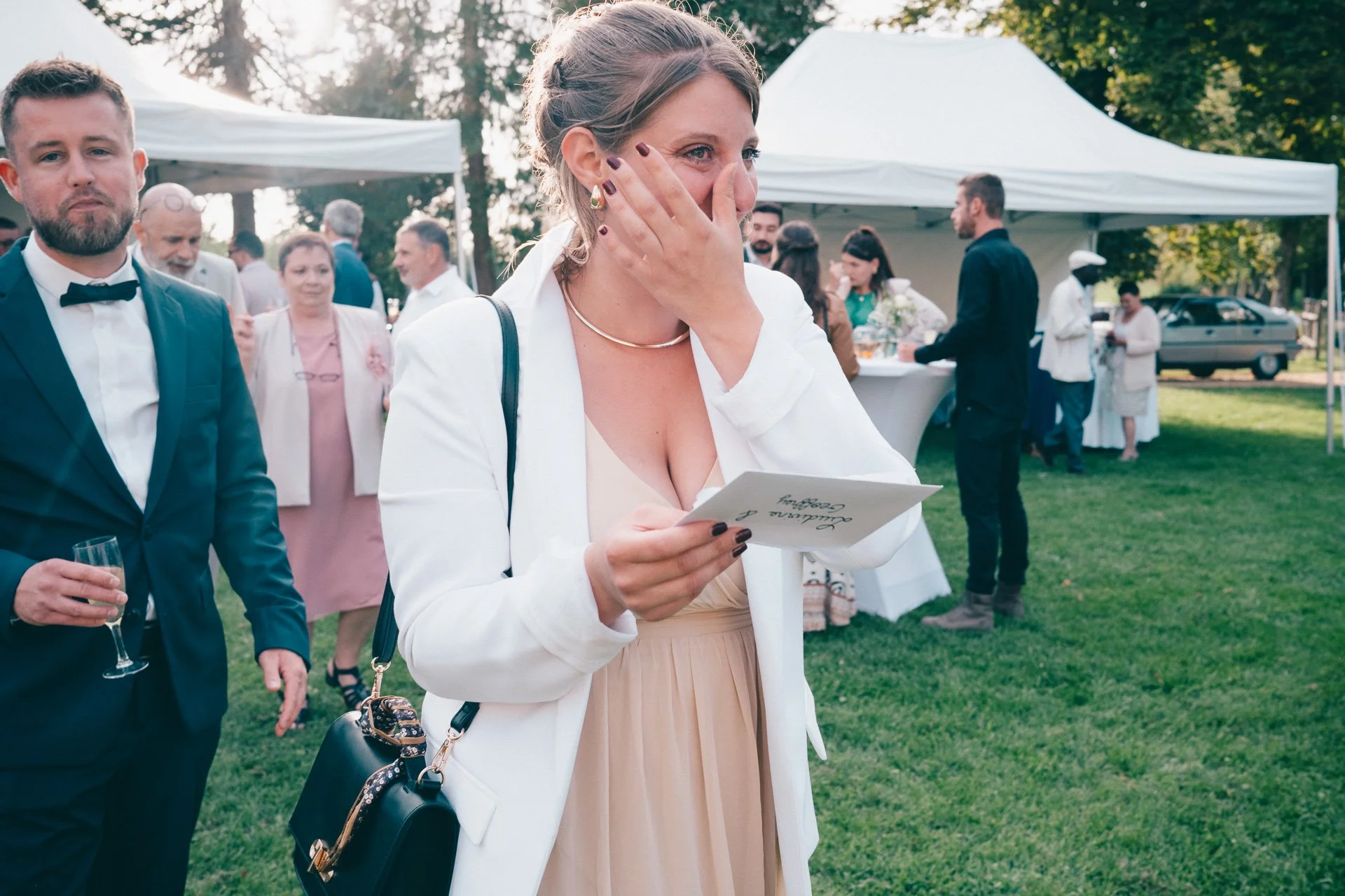 Une femme souriante portant une robe beige et une veste blanche, tenant une invitation ou un programme, lors d'une célébration ou d'un mariage en extérieur, entourée de personnes élégantes dans un cadre verdoyant avec des tentes.