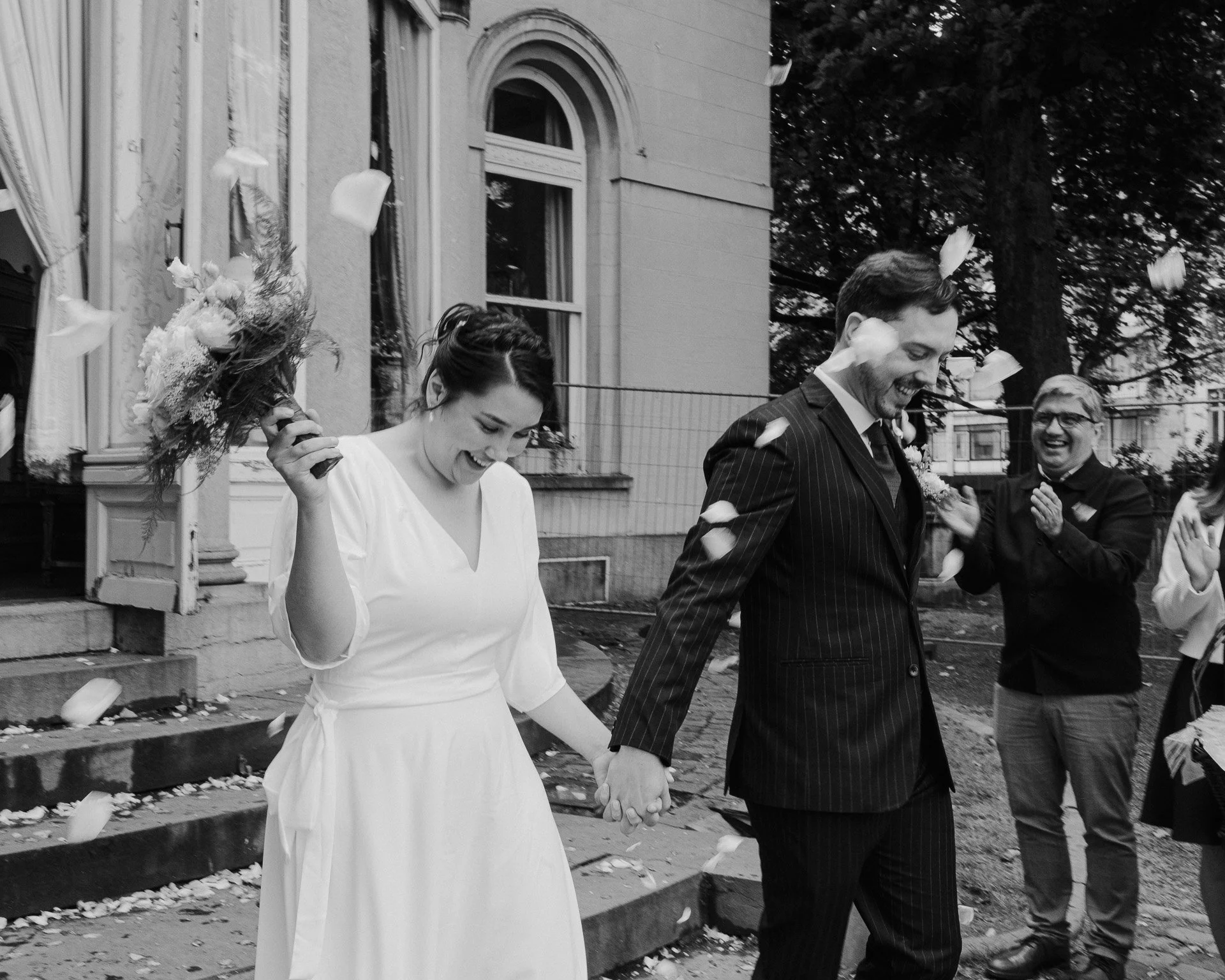 Un couple marié marche main dans la main lors d'un mariage en extérieur, avec des amis ou famille qui applaudissent derrière eux. La scène est en noir et blanc.
