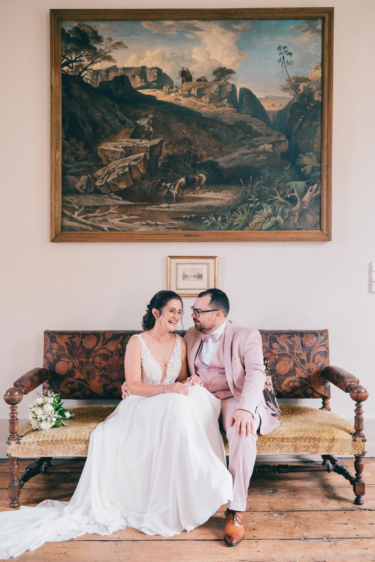 Un couple souriant habillé en mariage, assis sur un canapé en bois antique avec un cadre de tableau derrière eux.