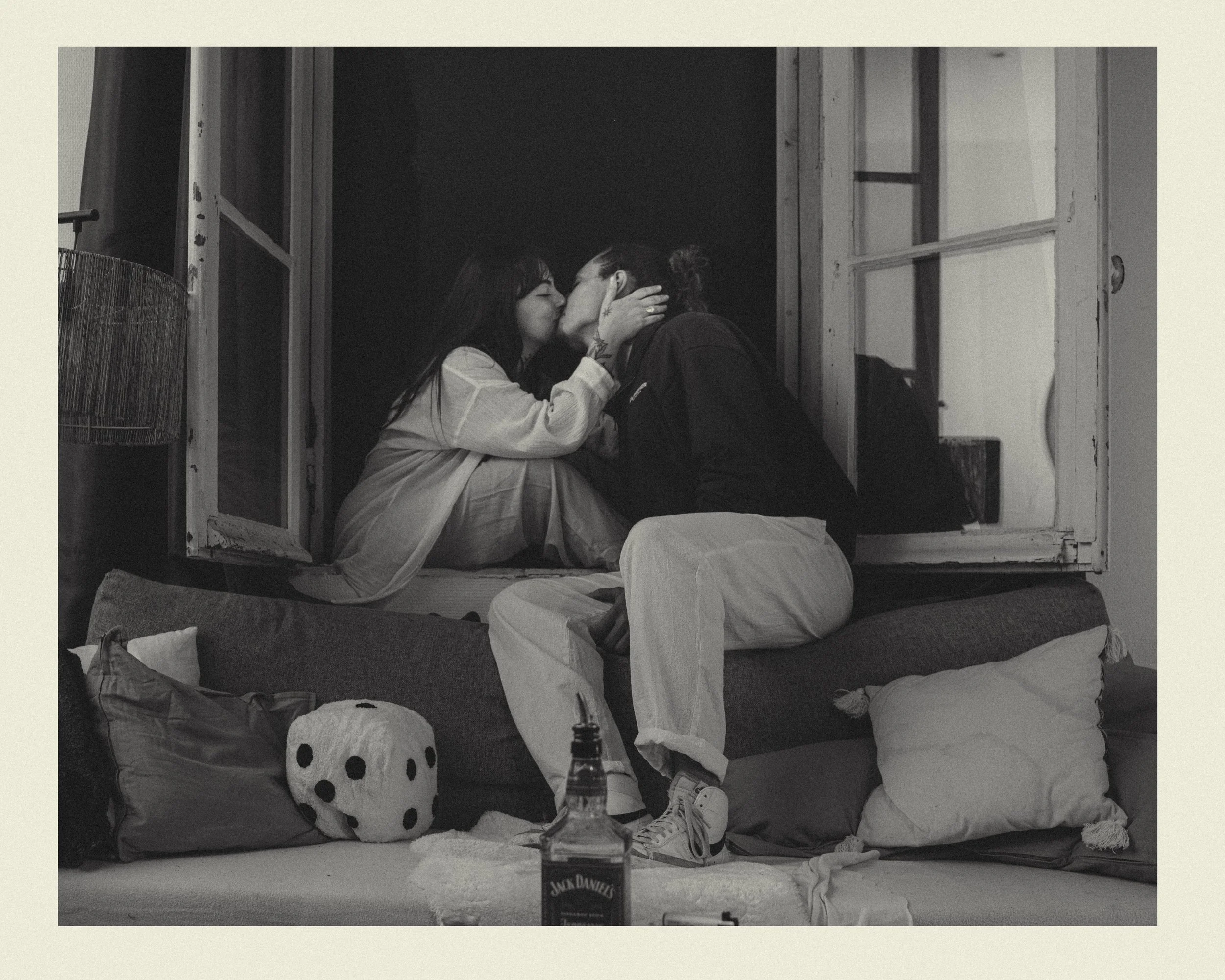Deux personnes, une femme et un homme, s'embrassent tendrement dans une pièce sombre, près d'une fenêtre ouverte. Autour d'eux, il y a des coussins, un ami avec des oreilles de polochon, une bouteille d'alcool dans une main, et un dés en forme de bou
