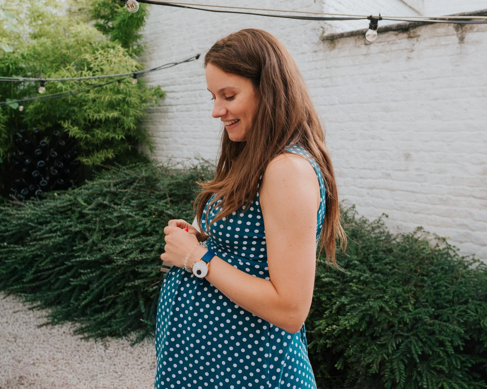 Femme aux cheveux longs, portant une robe bleue à pois blancs, souriant et regardant vers le bas, debout dans un jardin avec des plantes et une clôture blanche.