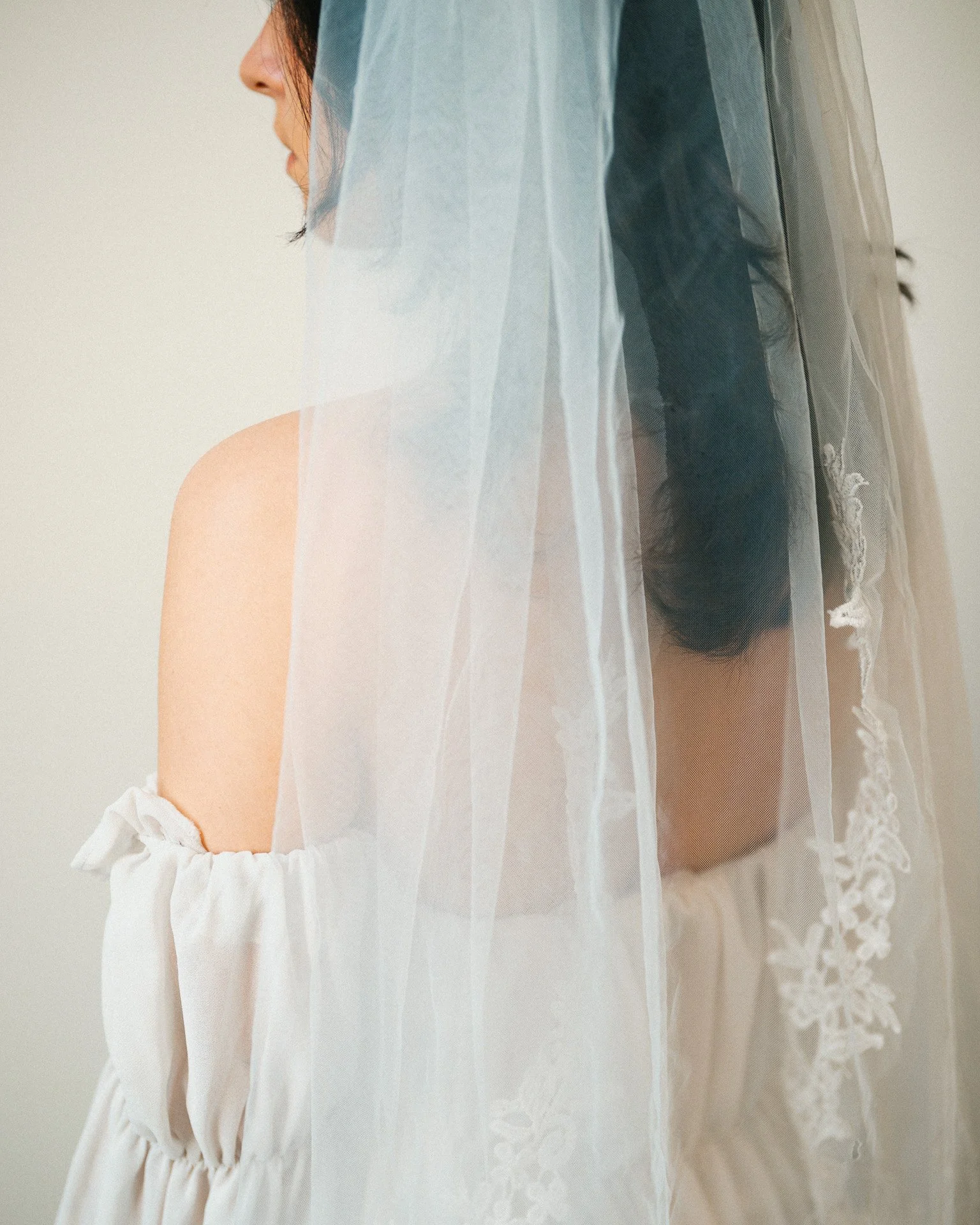 Une femme, vue de profil, portant une robe blanche avec un ruban près du bras, derrière une voile en tulle.