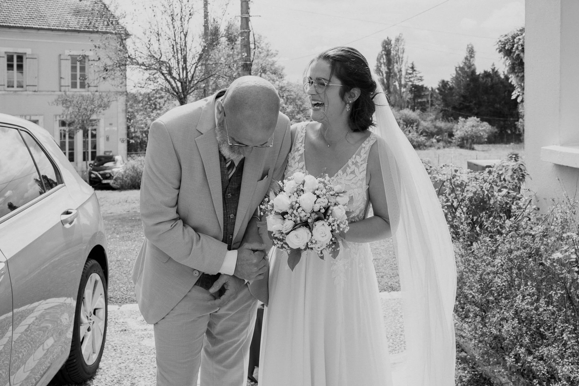 Une mariée souriante en robe blanche tient un bouquet de fleurs, accompagnée d'un homme plus âgé en costume conçu probablement pour la cérémonie impériale. Ils se tiennent la main, semble-t-il, dans une atmosphère de bonheur lors d'un mariage en exté