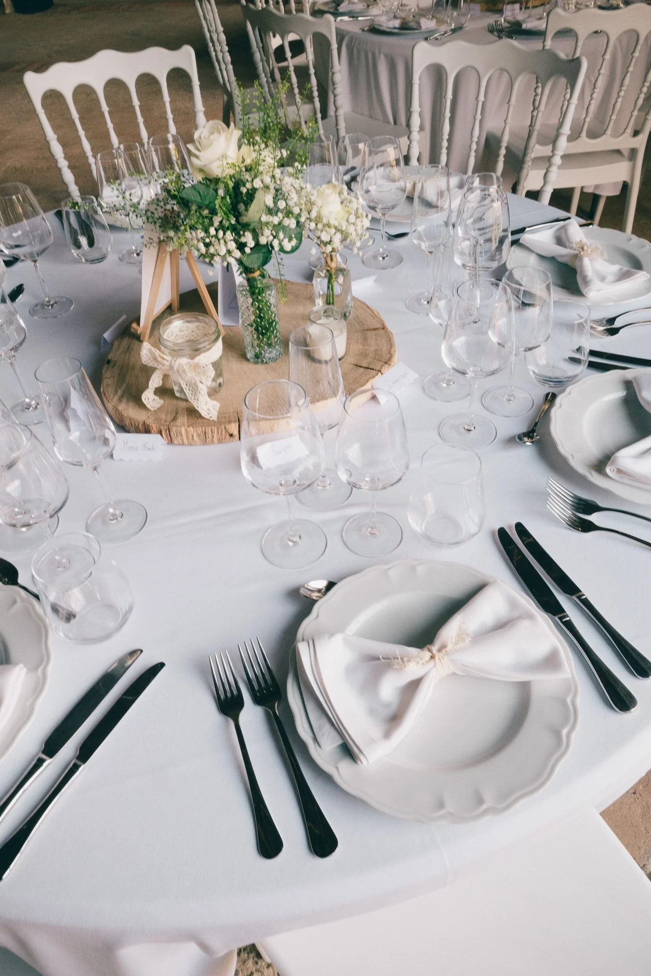 Une table de réception élégante avec une décoration florale centrale, des assiettes en porcelaine blanche, des verres à vin, des couverts en acier inoxydable, des serviettes pliées avec un ruban, et une nappe blanche.
