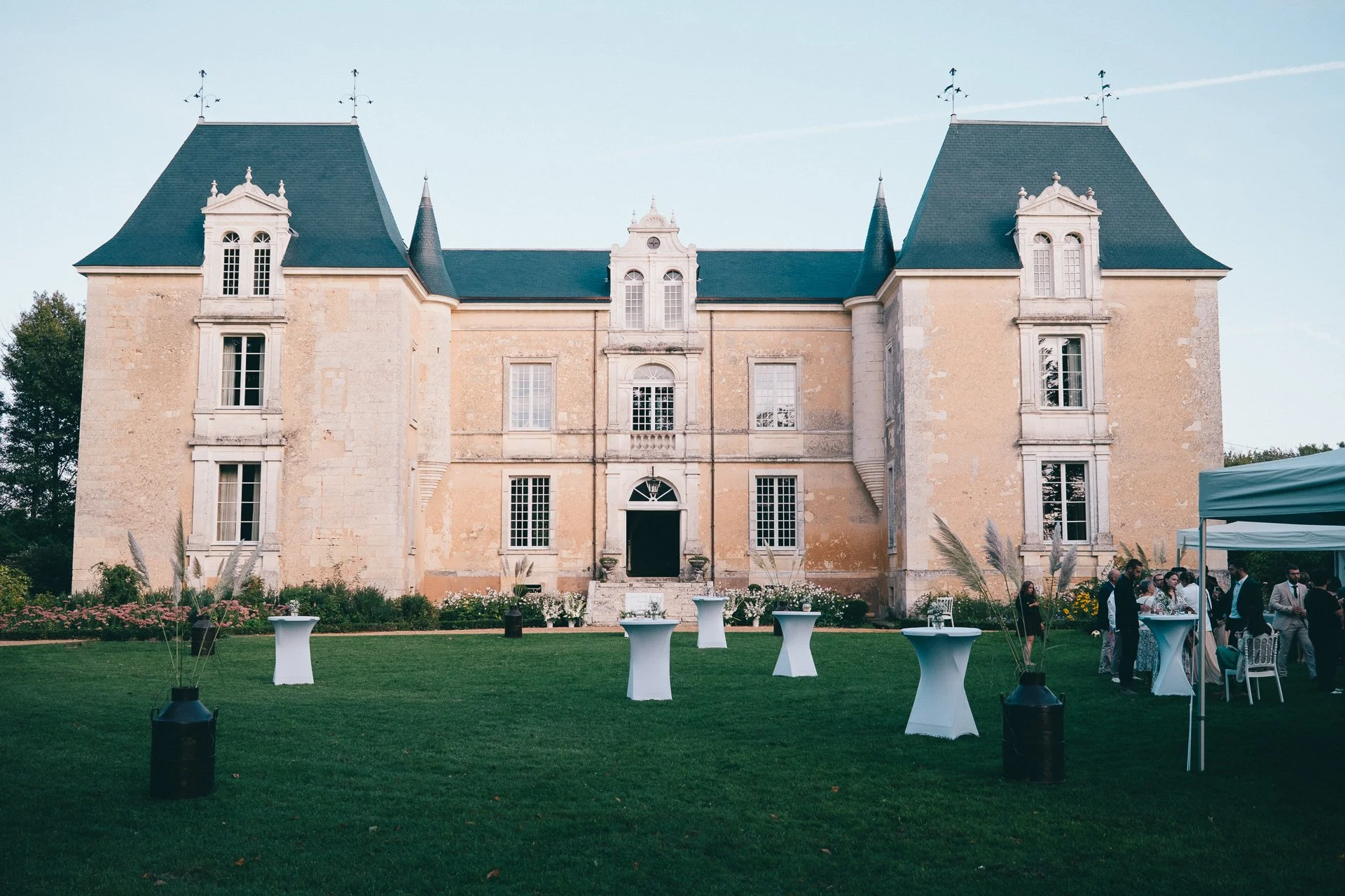 Un grand château en pierre avec un toit bleu, entouré d’un jardin bien entretenu, lors d’un événement en plein air avec des groupes de personnes et des tables hautes blanc.