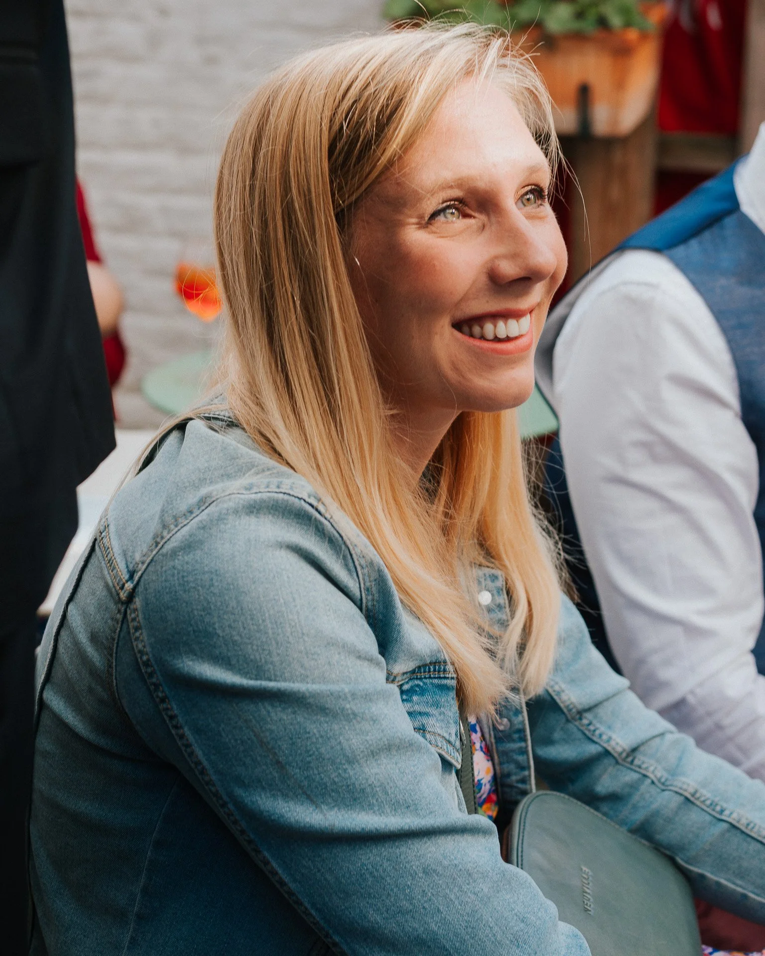 Une femme aux cheveux blonds souriante assise lors d'une réunion ou événement social, portant un jean. Personne à côté d'elle, décor avec plantes en arrière-plan.