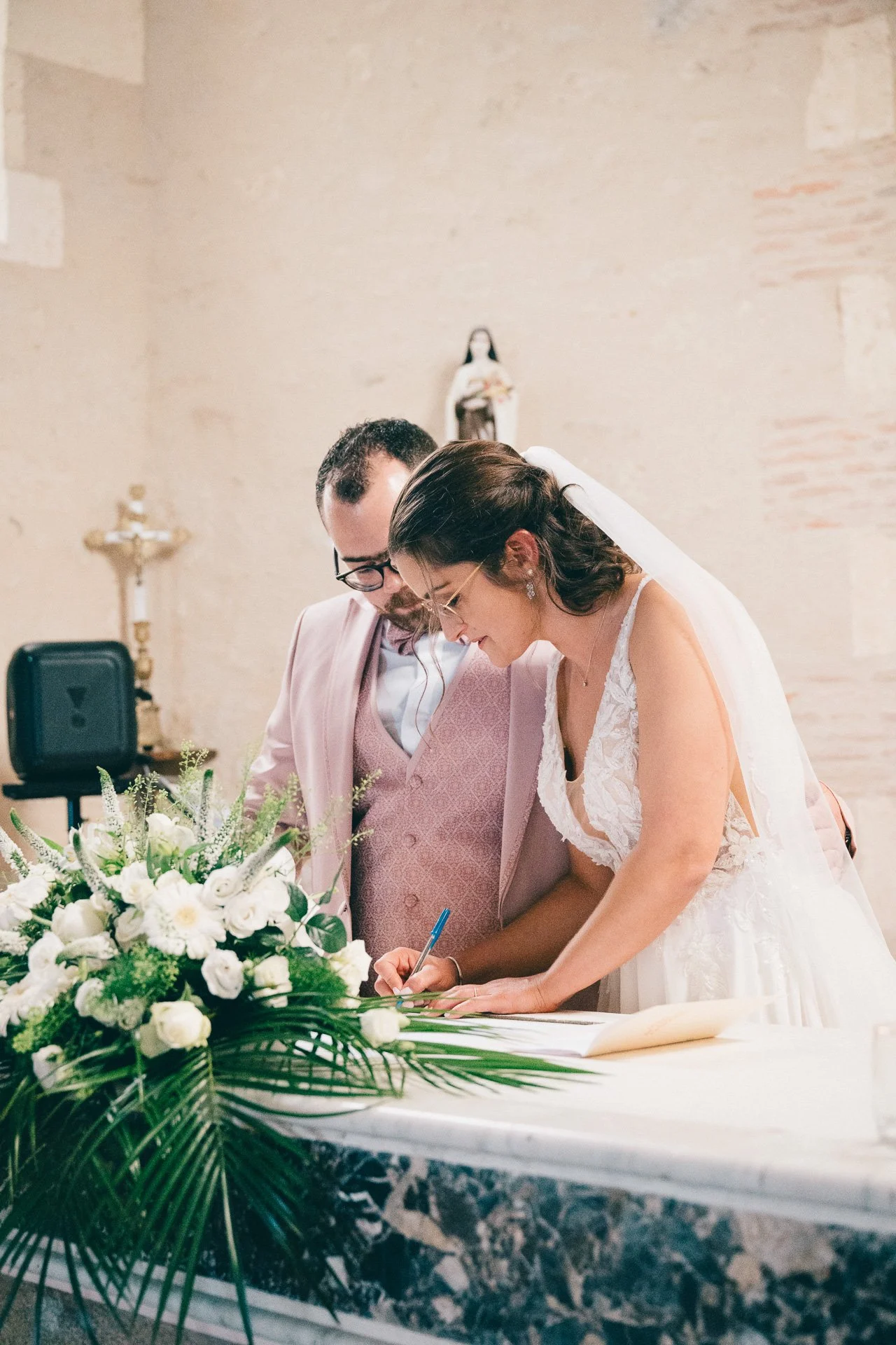 Un couple de mariés signe un registre de mariage dans une église, entouré de fleurs blanches et de décorations religieuses.