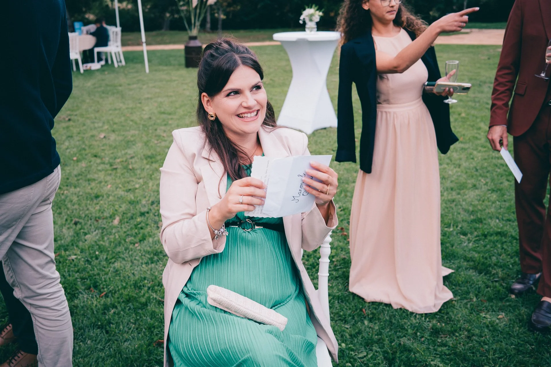 Une femme souriante, habillée en vert et beige, assise sur une chaise lors d'une fête en plein air, tenant une carte ou une enveloppe, entourée d'autres personnes en conversation.