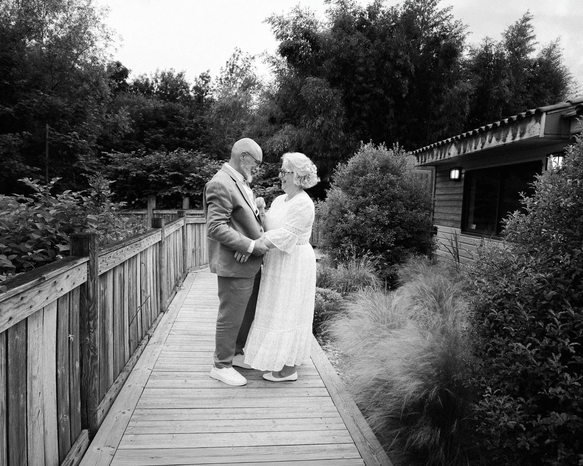 Un couple âgé se tient la main sur un pont en bois dans un jardin, échangeant un regard affectueux, avec une maison en bois et des arbres en arrière-plan.