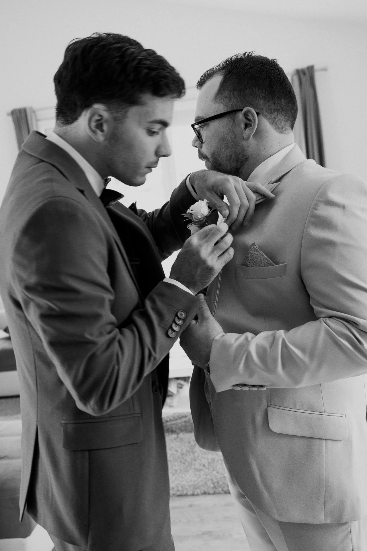 Deux hommes en costard, l'un aidant l'autre à boutonner son veston, lors d'une cérémonie ou d'une occasion spéciale.