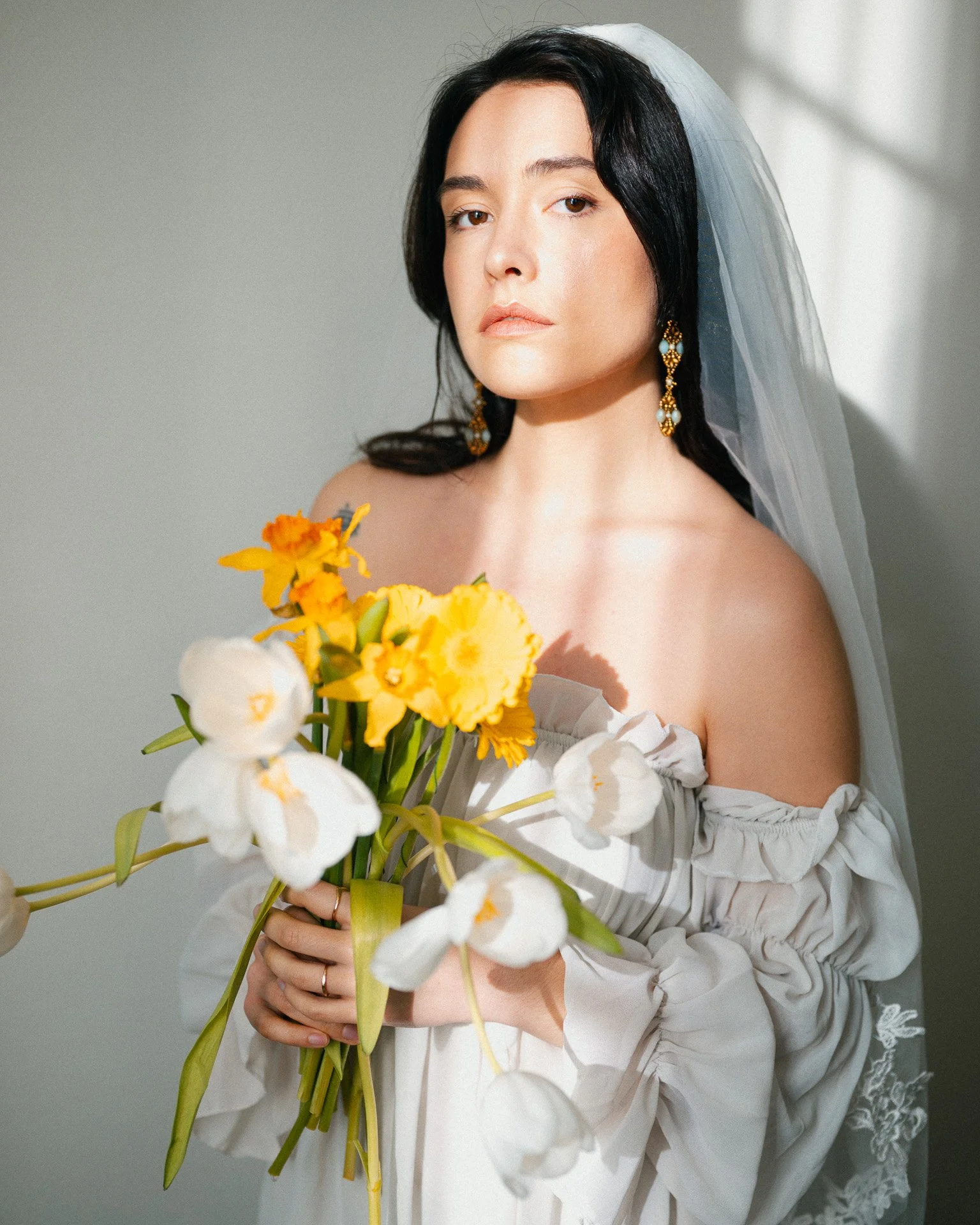 Une femme portant une robe blanche, une voilette, et de grosses boucles d'oreilles, tenant un bouquet de fleurs blanches et jaunes.