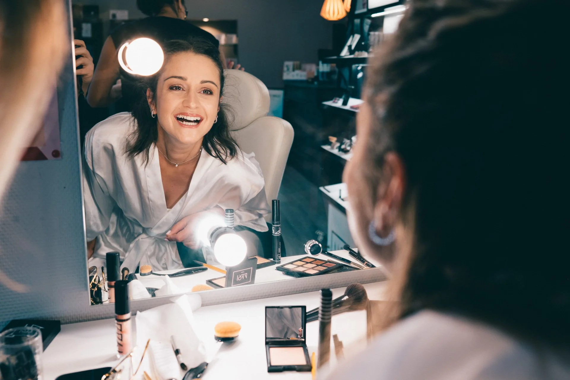 Femme souriante regardant son reflet dans un miroir, entourée de maquillage et d'outils de beauté, dans un espace de maquillage ou de studio.