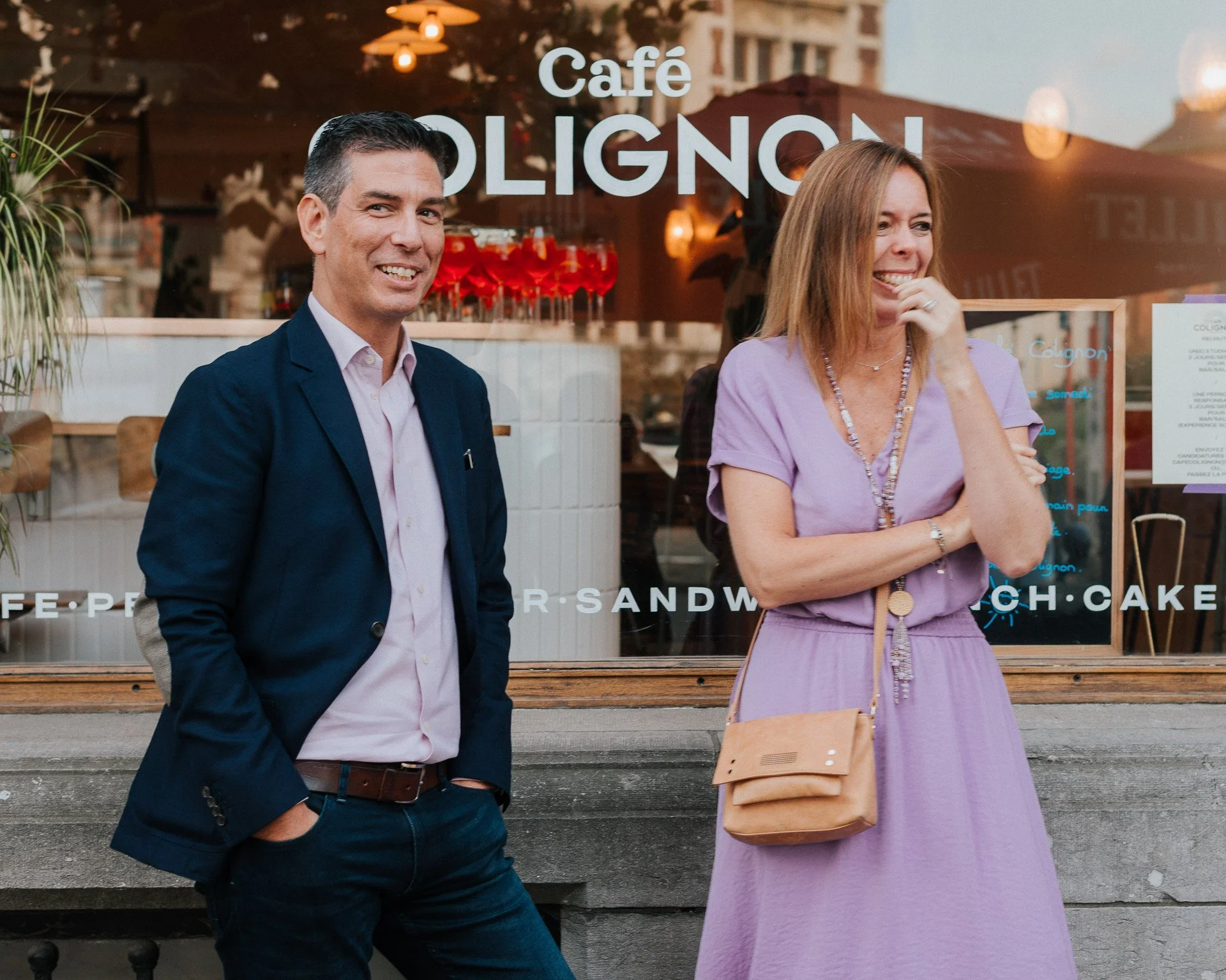 Deux personnes, un homme en costume bleu et une femme en robe violette, rient et discutent devant une vitrine d'un café à l'extérieur.