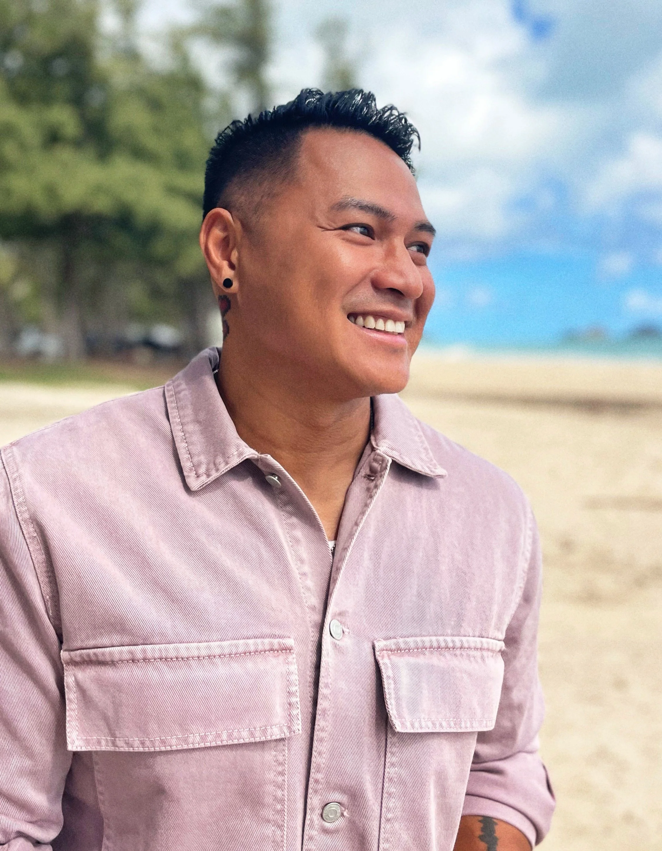 Close-up of a smiling man with styled hair, black earrings, and tattoos on his neck and arm, wearing a light purple denim jacket, outdoors with trees and a beach in the background.