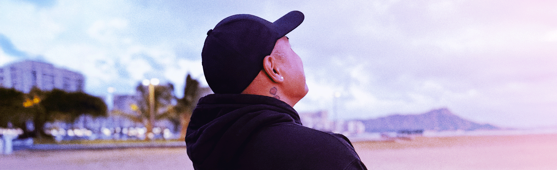 A person wearing a black cap and hoodie standing on a beach, looking at the ocean with a cityscape and mountains in the background.