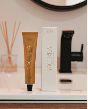 A tube of ALGENA skincare product and its white box packaging on a bathroom counter, with a mirror, black faucet, and reed diffuser in the background.