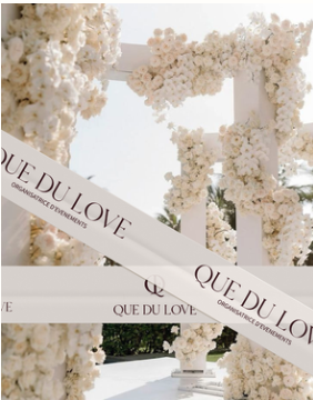 Wedding arch decorated with white flowers with a ribbon that says 'QUE DU LOVE' across it.