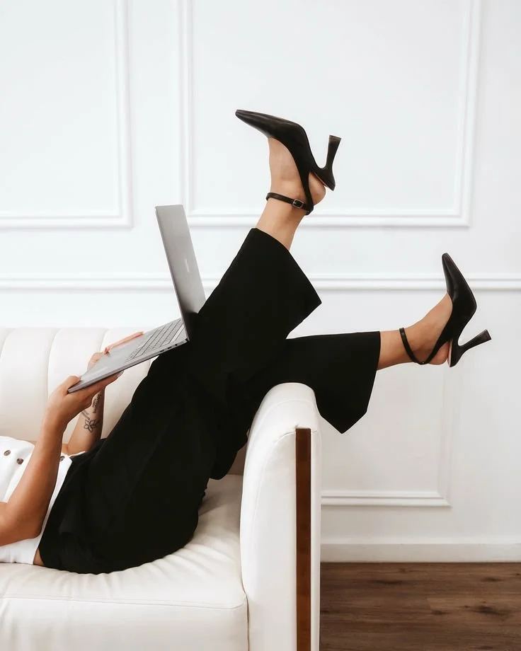 Person reclining on a white sofa with legs raised and crossed, wearing black high heels and using a laptop.