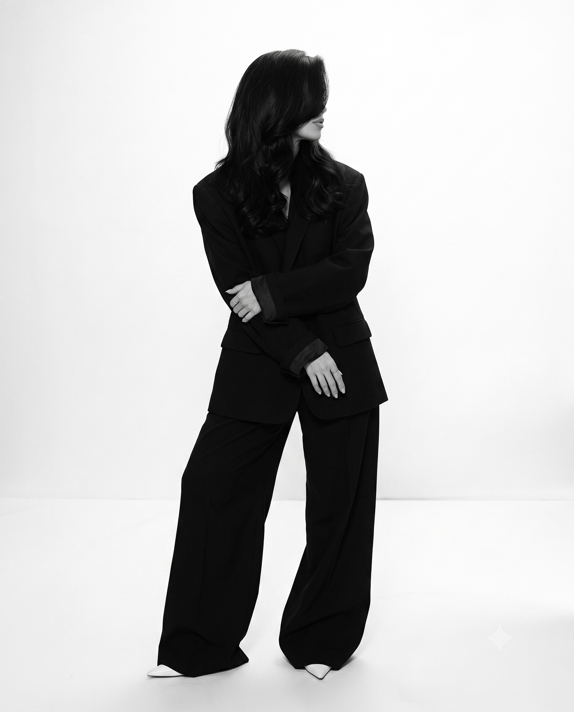 A woman with long wavy black hair wearing a black suit and black wide-leg pants, standing against a plain white background, with her arms crossed and looking to her right.