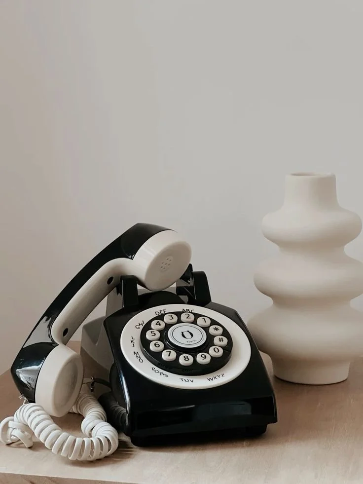 A vintage rotary phone in black and white with a curved handset, placed on a light-colored surface next to a white ceramic sculpture with tiered, organic shapes.