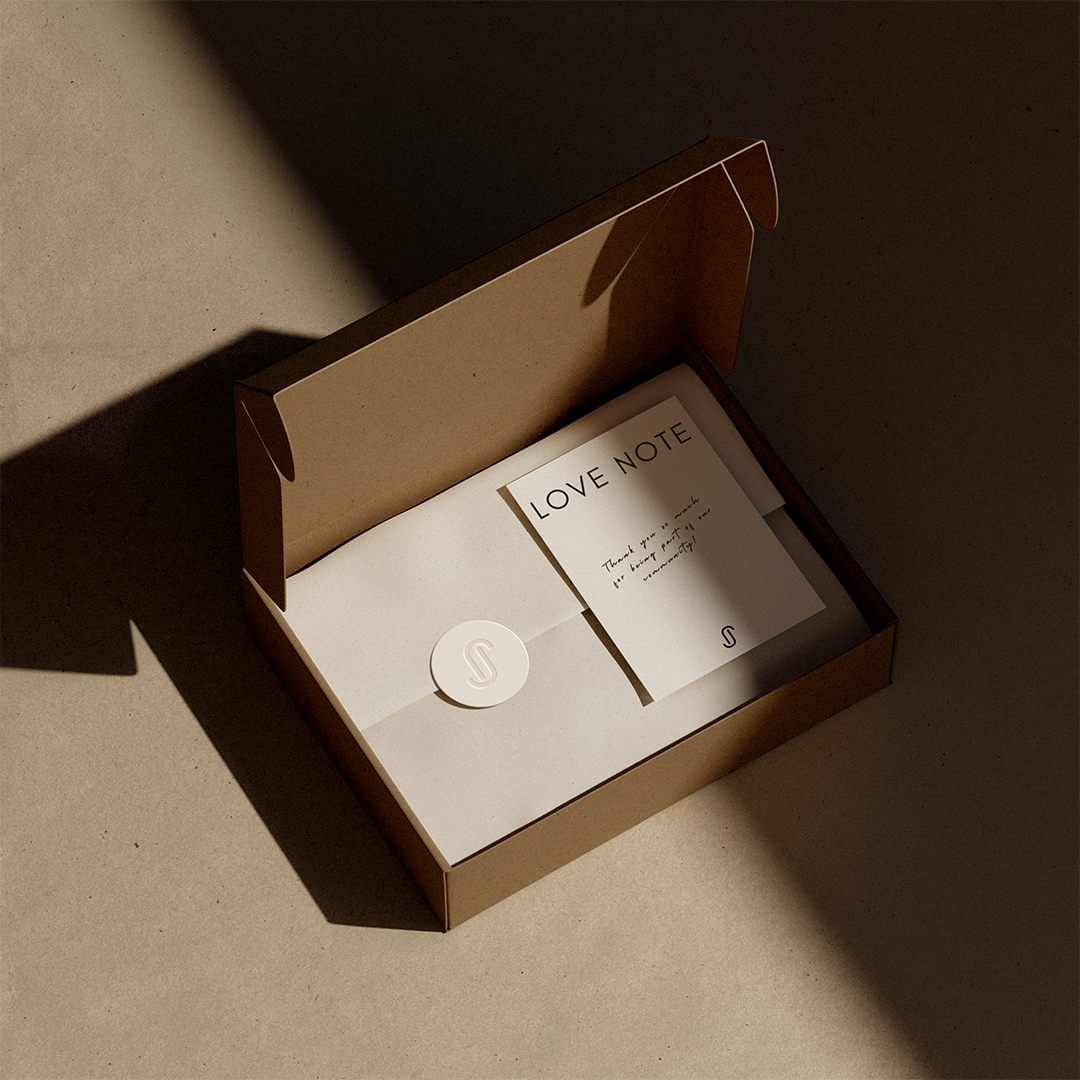 An open cardboard box containing a note labeled "Love Note" and an envelope with a seal, placed on a textured beige surface with shadows.