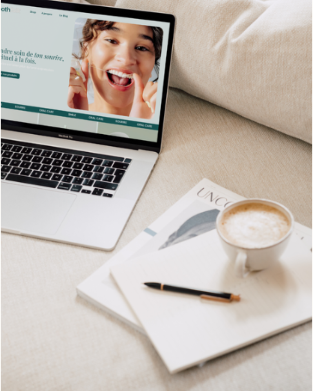 A MacBook displaying a woman smiling and brushing her teeth, placed on a beige fabric surface with a coffee cup, pens, and magazines nearby.