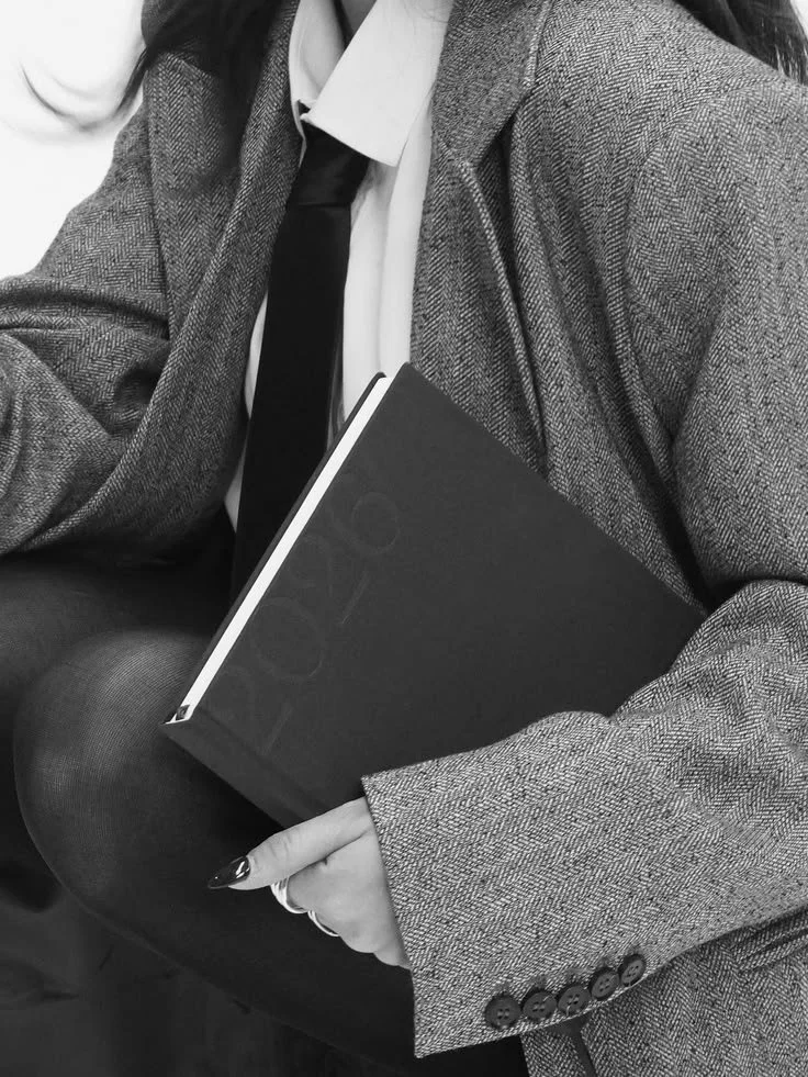 A woman wearing a blazer, white shirt, and tie, holding a closed notebook or journal with a dark cover.