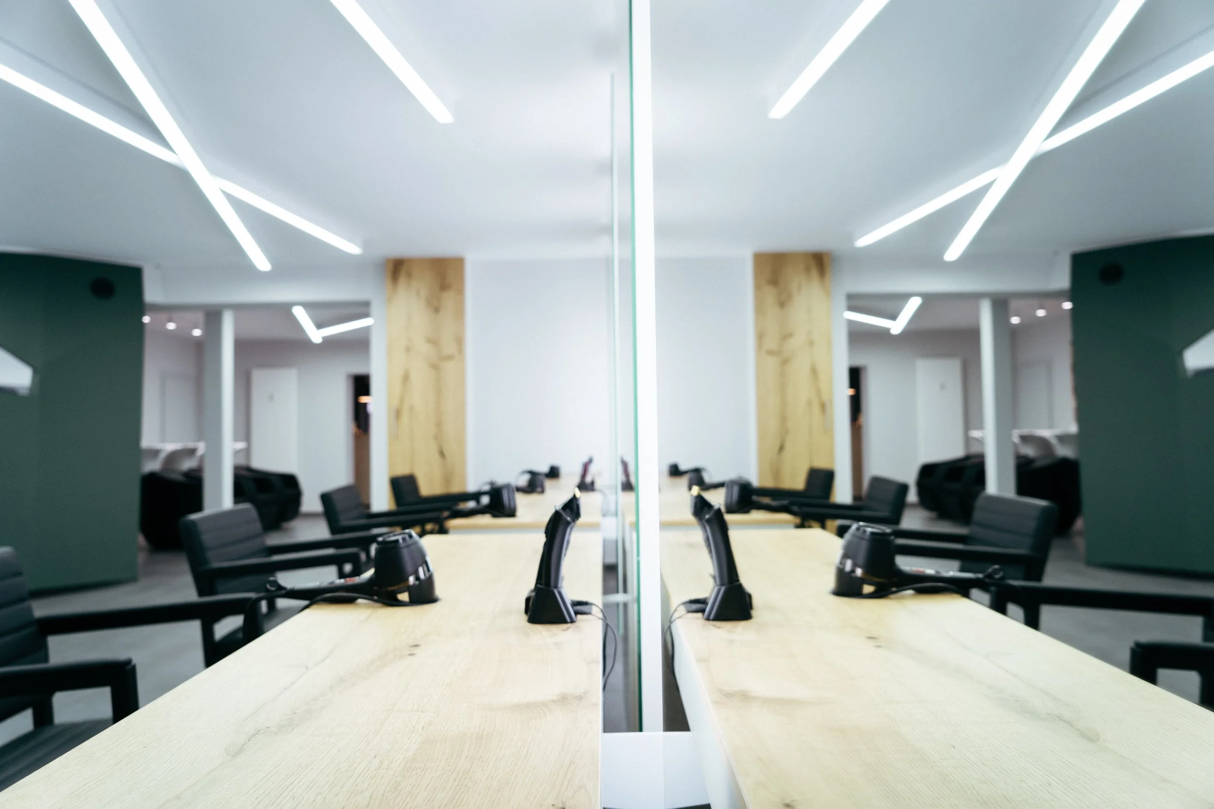 Ein moderner Besprechungsraum mit einem langen Tisch, schwarzen ergonomischen Stühlen und einer reflektierenden Glasscheibe in der Mitte. Die Decke ist mit weißen Lichtleisten beleuchtet, Wände mit Holzakzenten und dunklen Wandflächen. Im Hintergrund
