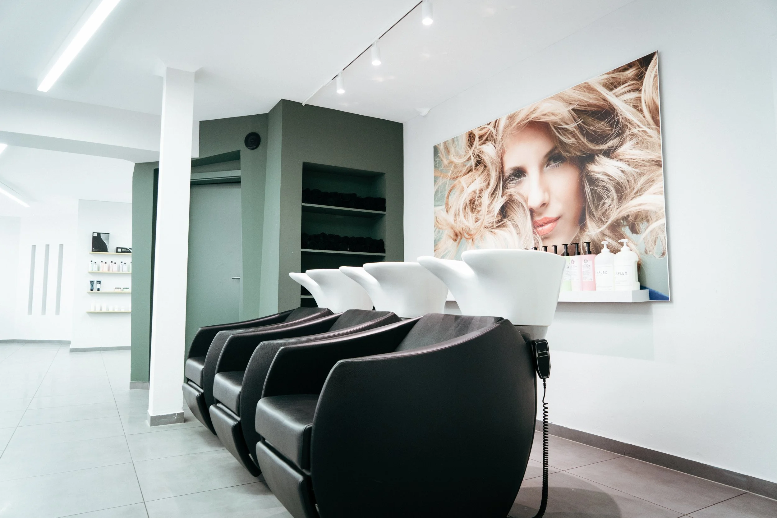 Ein Friseursalon mit schwarzen Waschbecken, weißen Stühlen, einem großen Portrait einer Frau mit lockigem blondem Haar an der Wand und Regalen mit Pflegeprodukten.