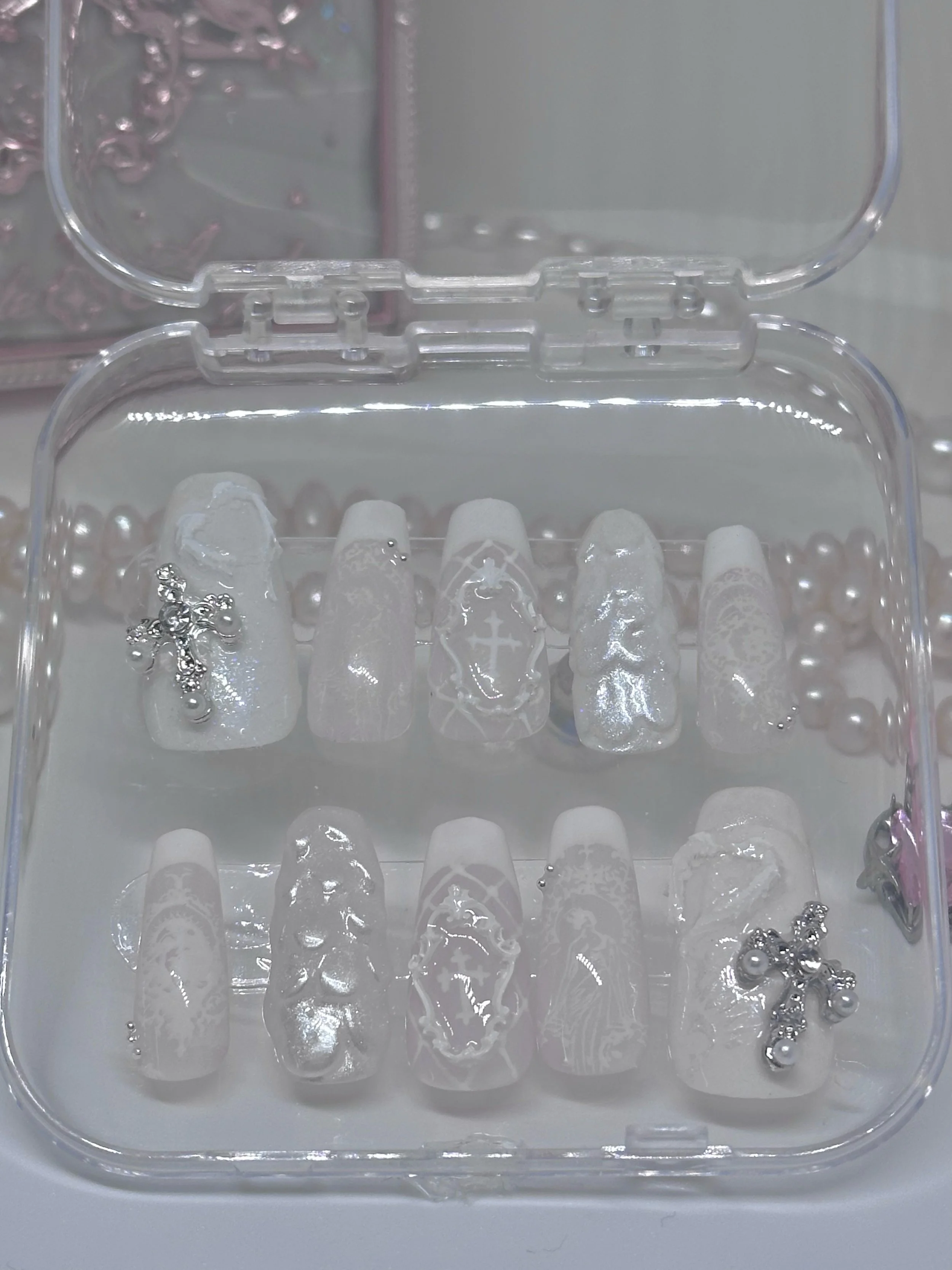 Clear plastic container holding decorative clear glass nails with white snowflake and cross patterns, some small silver beads