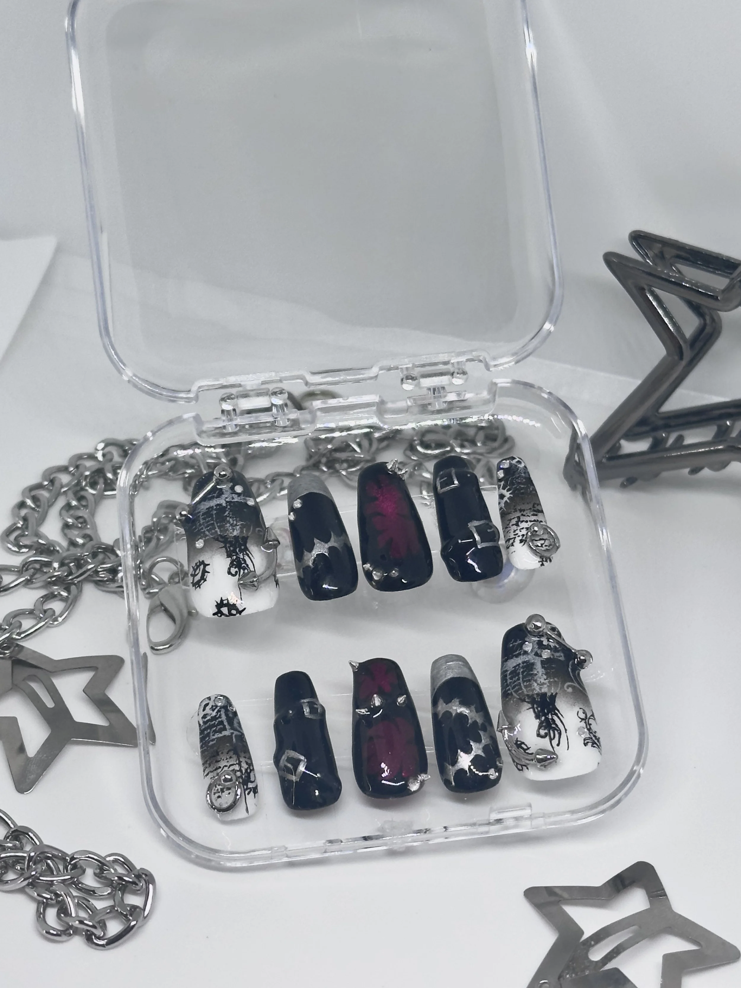 Display case of assorted nail art press-on nails with various black, white, and pink designs, alongside metal jewelry chains and star-shaped clips on a white surface.