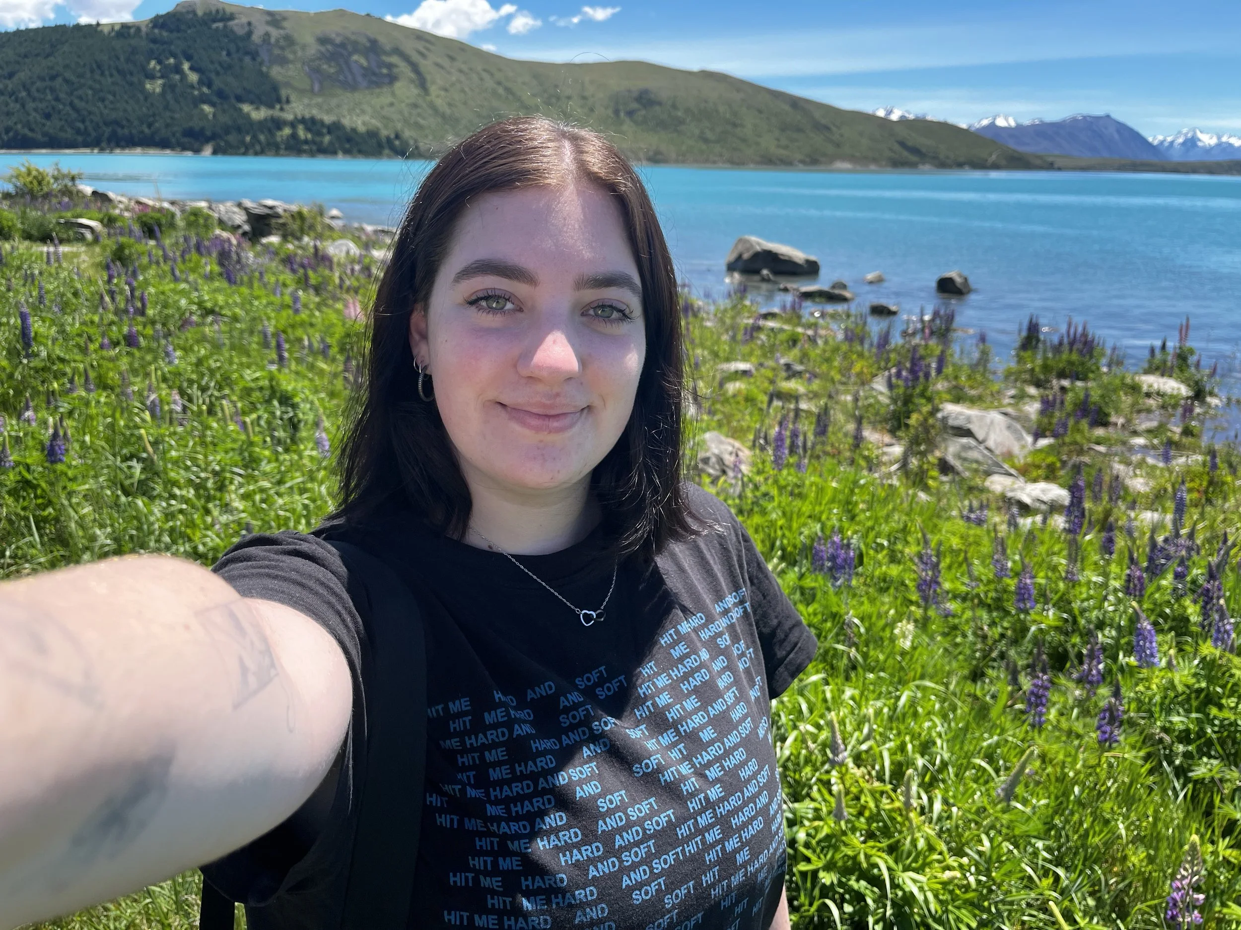 A young woman taking a selfie outdoors near a lake. The background features a calm, blue lake surrounded by green hills and mountains with snow on their peaks. The woman has long dark hair, light-colored eyes, and is wearing a black T-shirt with blue text.
