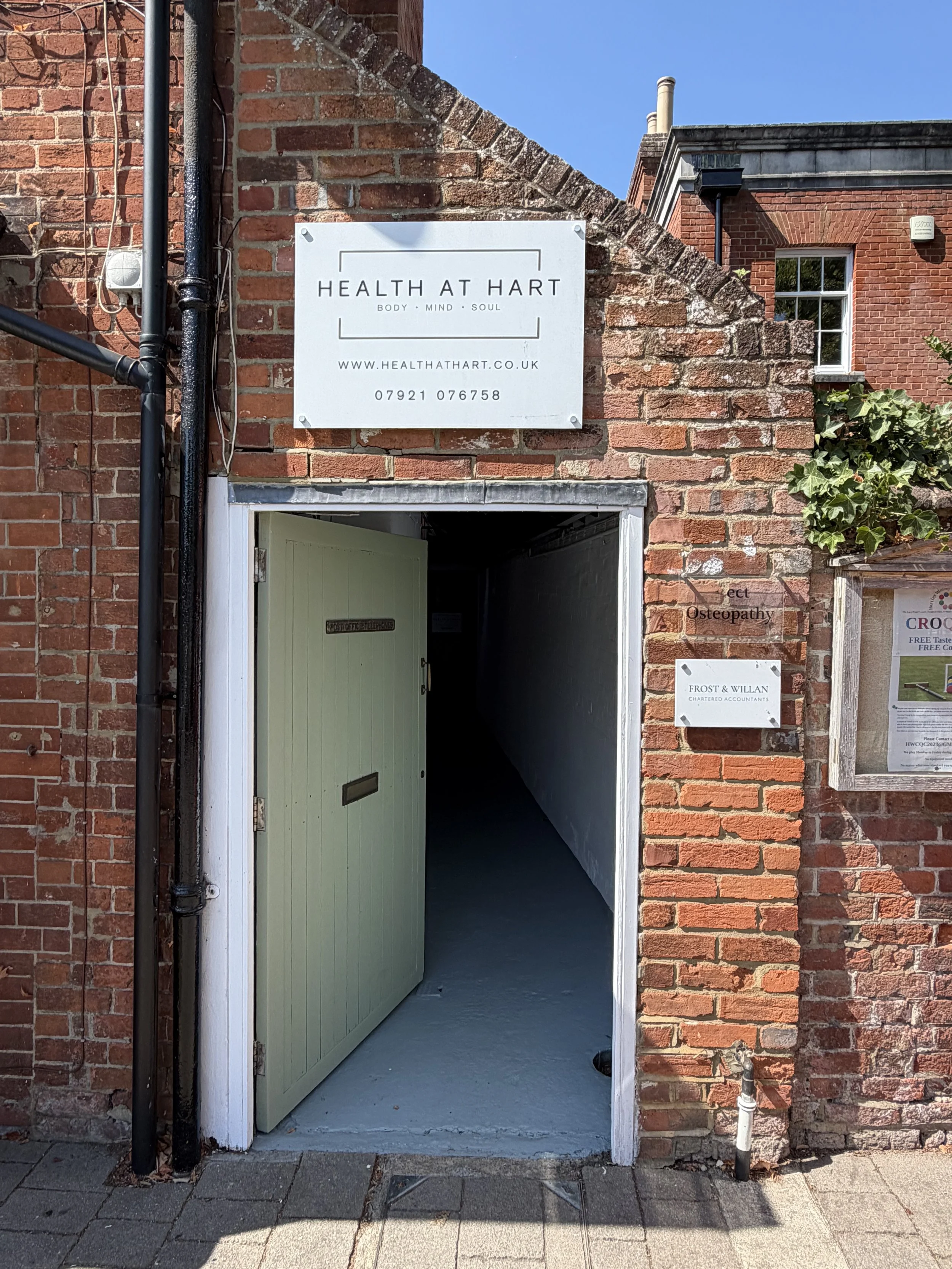 Open brick doorway with a pale green door slightly ajar, leading into a dimly lit corridor. Sign above the entrance reads 'Health at Hart' with the tagline 'Body, Mind, Soul' and a website address. There is a small sign on the brick wall to the right mentioning osteopathy. Sunlight casts shadows on the brick wall and sidewalk.