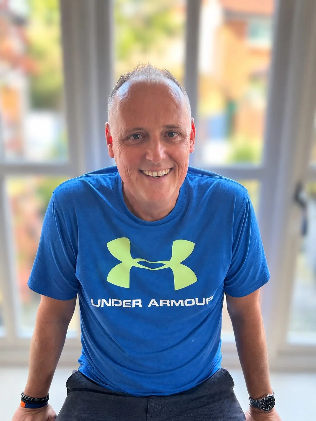 Man smiling in a blue Under Armour t-shirt standing in front of large windows with a blurred outdoor background.