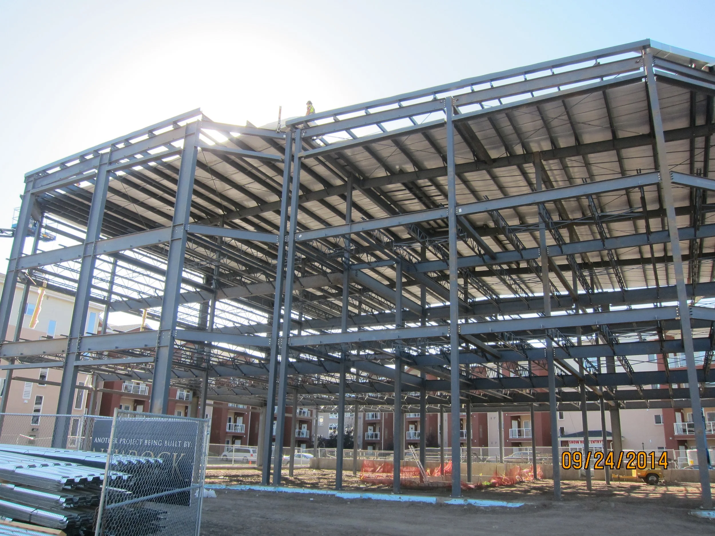Steel framework of a building under construction with multiple levels and a worker on top, set against a bright sky and residential apartment buildings in the background.