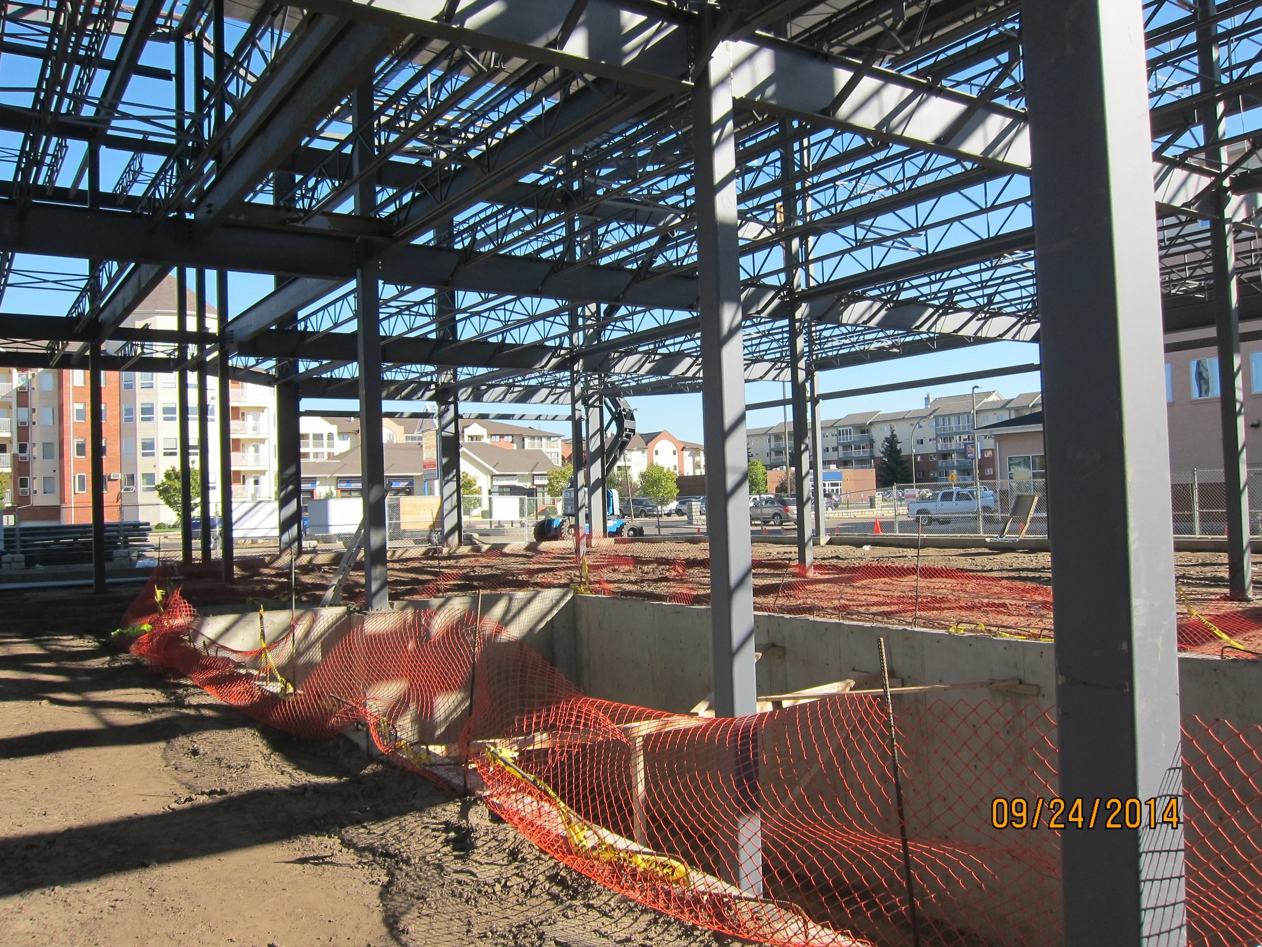 Construction site with steel framework and orange safety fencing, with multi-story apartment buildings in the background under a clear blue sky.