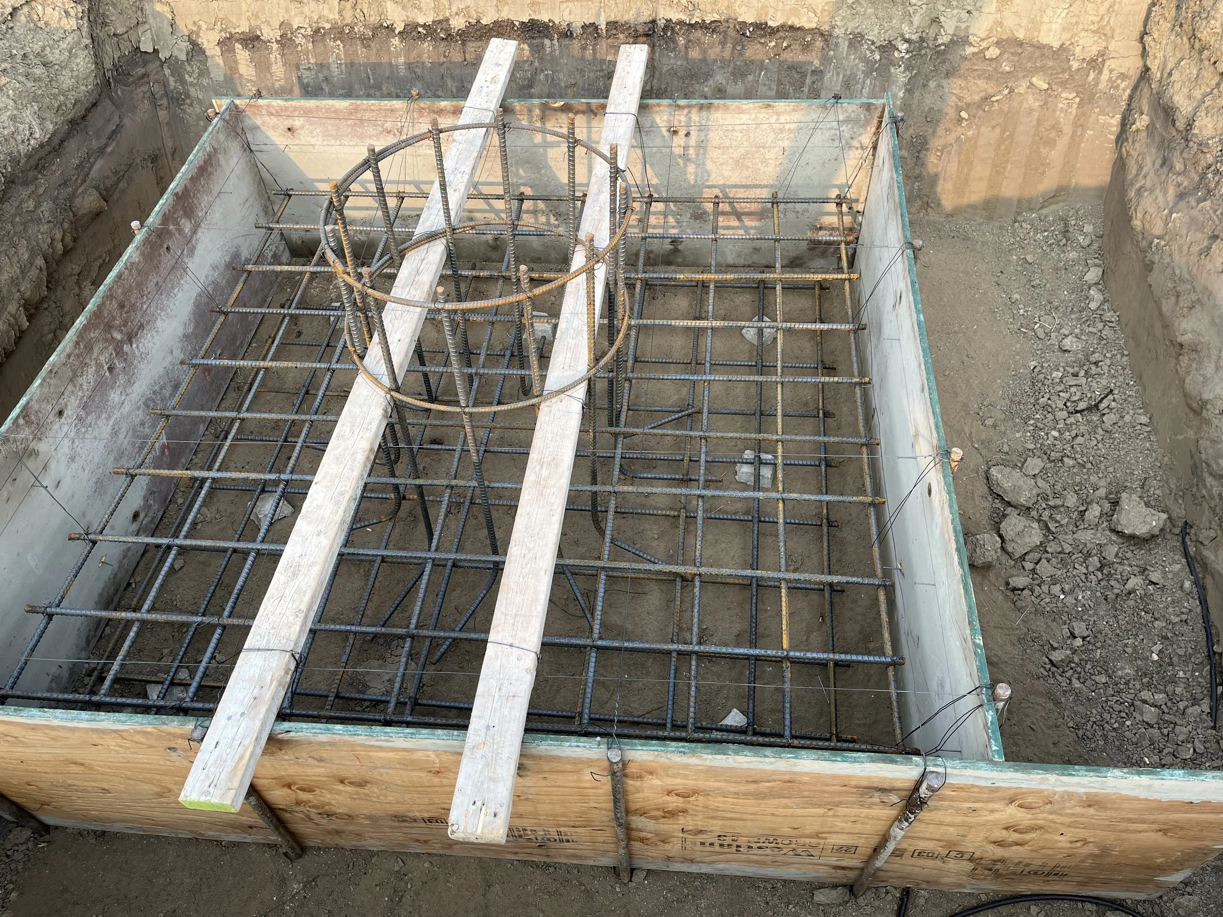 Construction site showing a concrete foundation with rebar reinforcement and wooden formwork, with two wooden planks laid across the rebar.