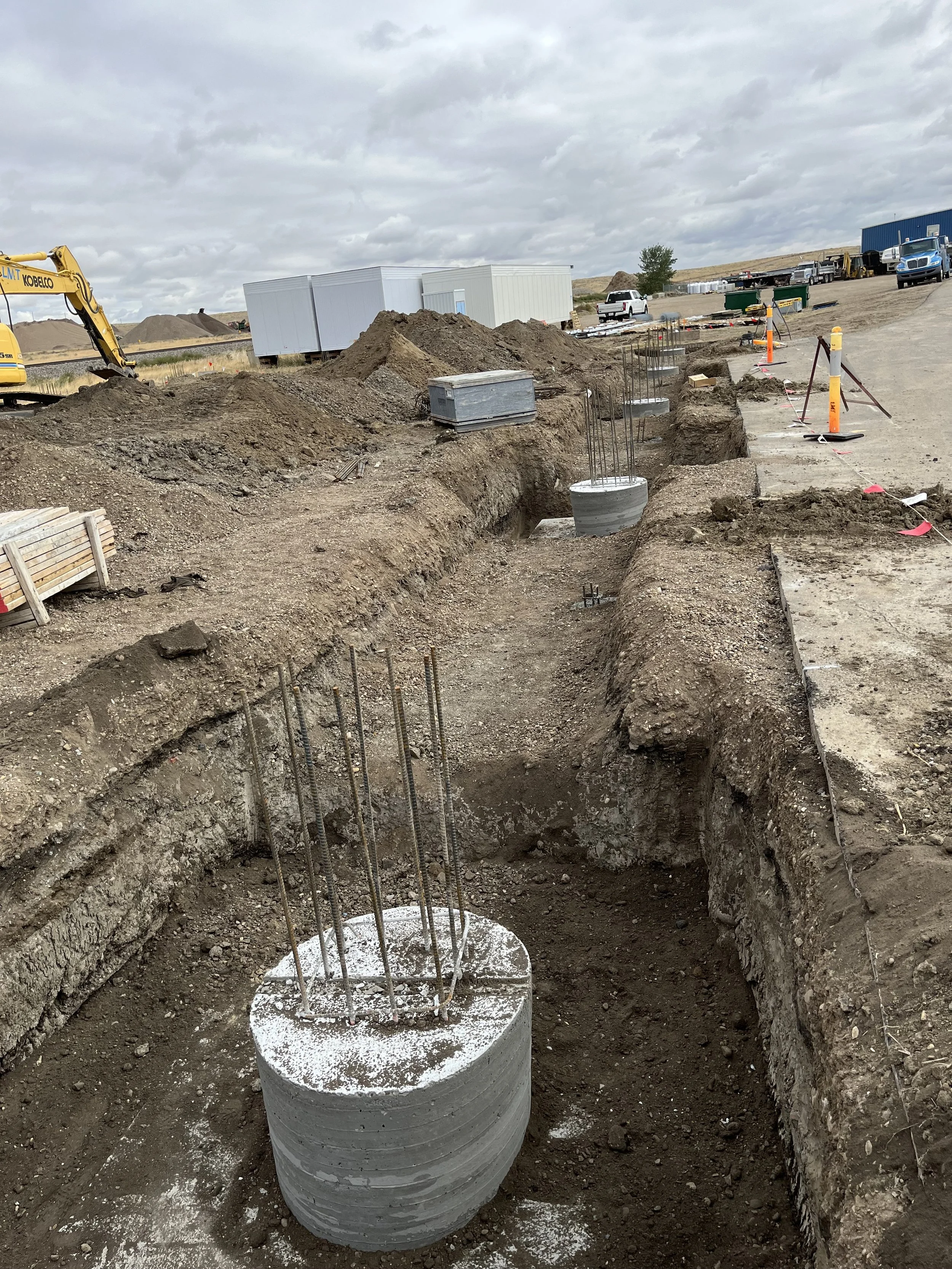 Construction site with concrete foundation and steel rebar for underground utilities, dirt piles, construction equipment, and vehicles under cloudy sky.