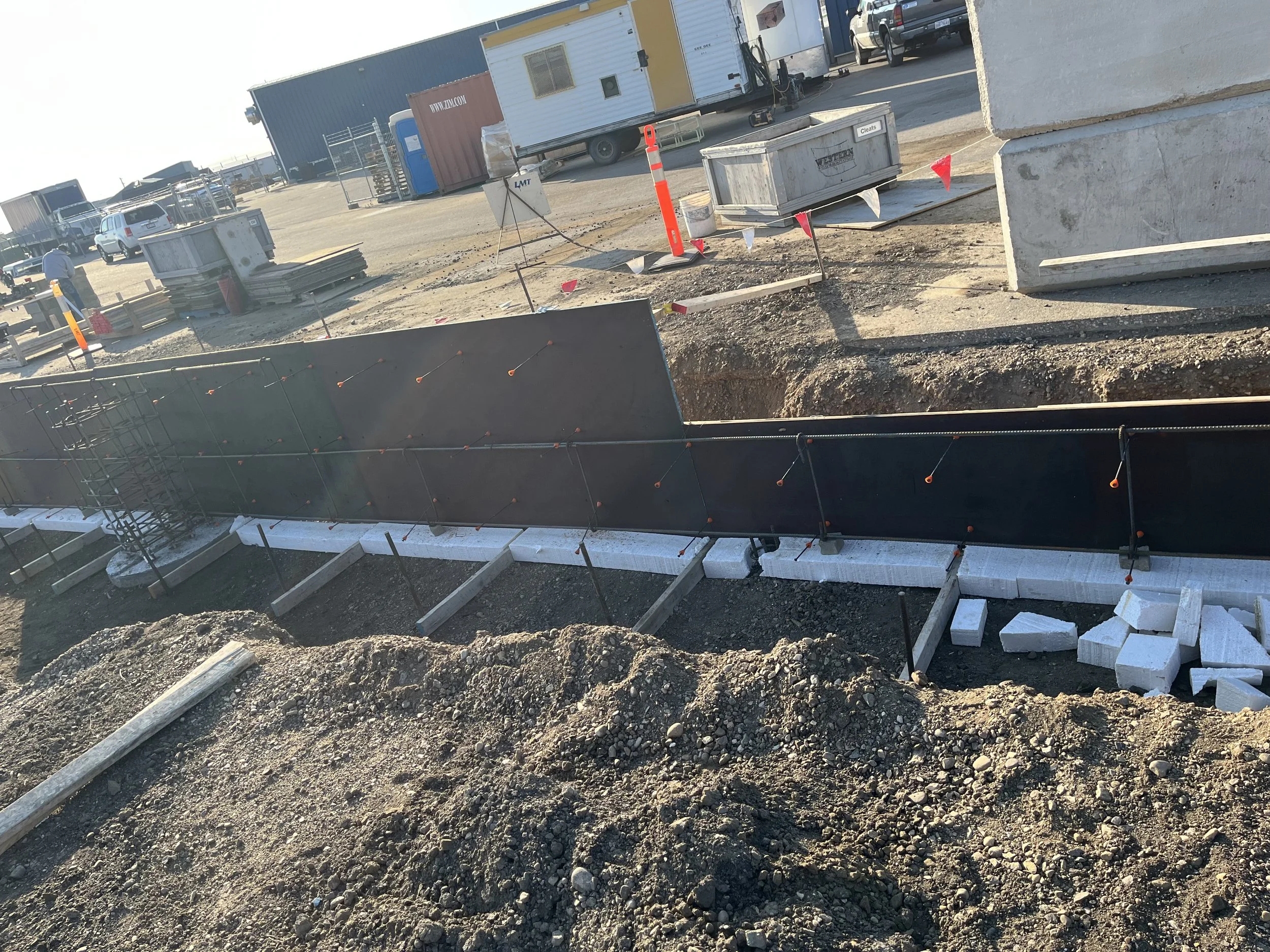 Construction site with dug-out area, concrete blocks, construction barriers, and equipment, with parked cars and trailers in the background.