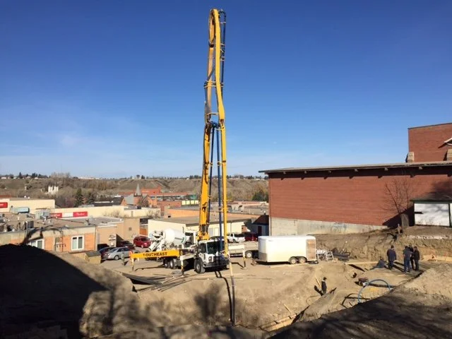 Construction site with a large yellow drilling machine next to a red brick building, with workers and construction equipment nearby.
