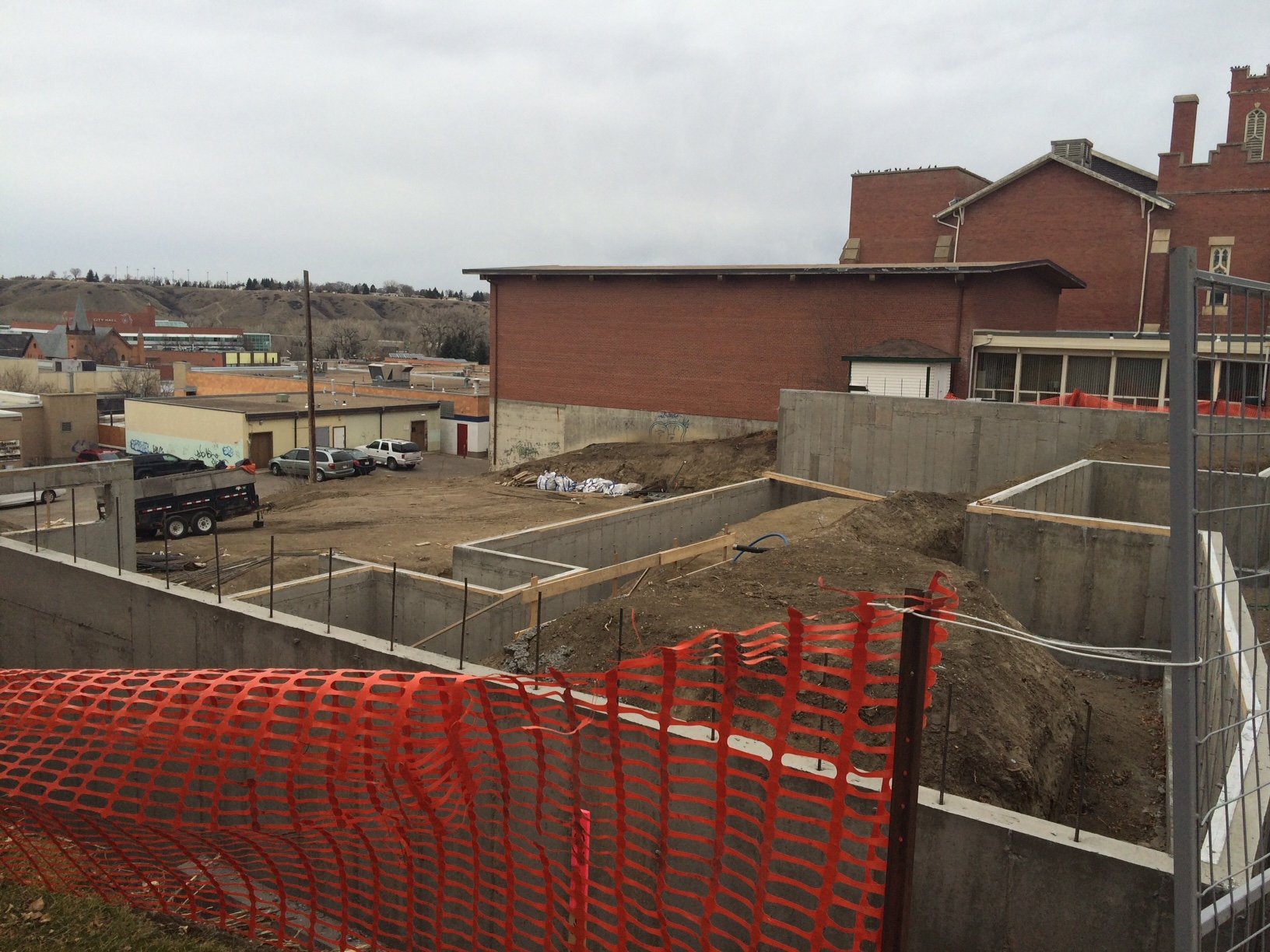 Construction site with dug foundation, orange safety fencing, and construction materials, surrounded by buildings and parking lot.