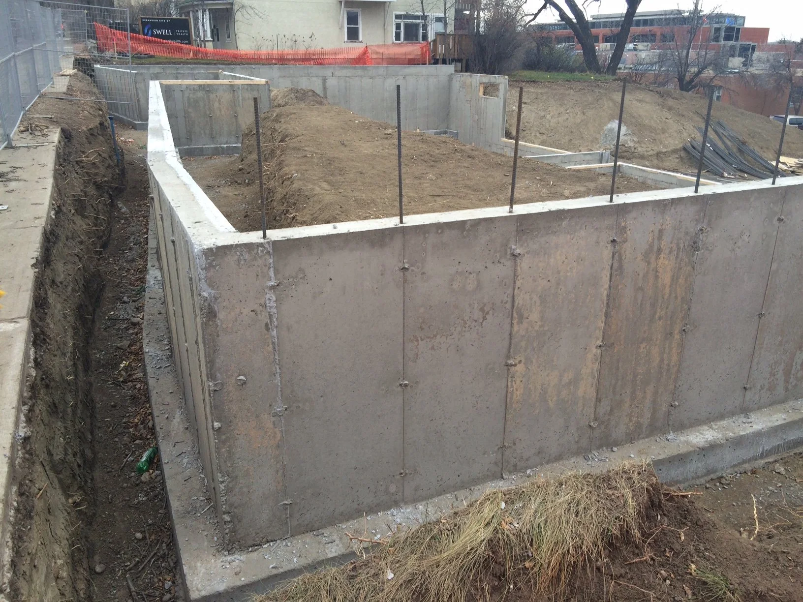 Construction site showing concrete foundation walls with rebar extending upward, surrounding a dirt excavation area.