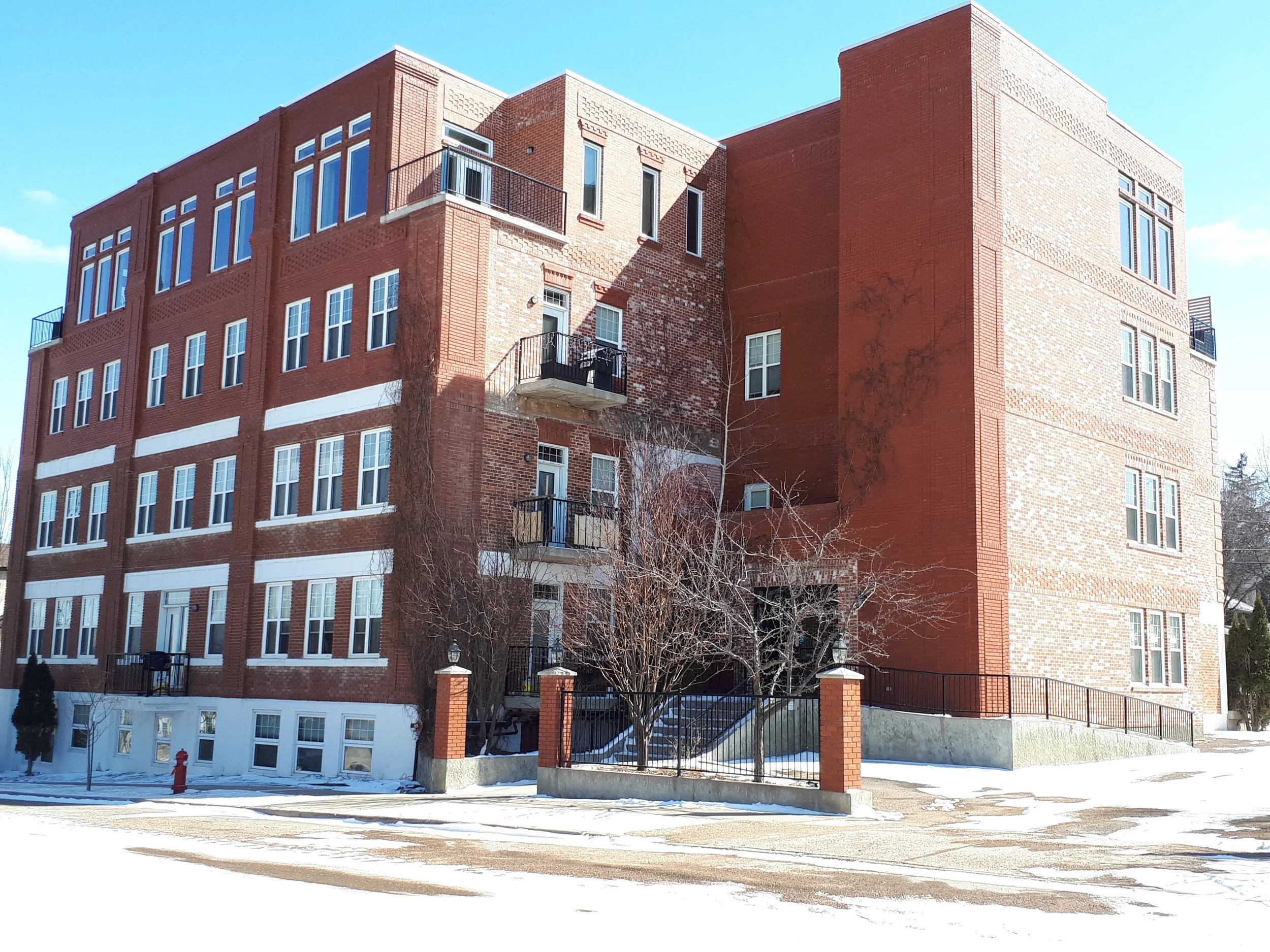 A multi-story brick apartment building with several windows, balconies, and a gated entrance, set against a clear blue sky with patches of snow on the ground.